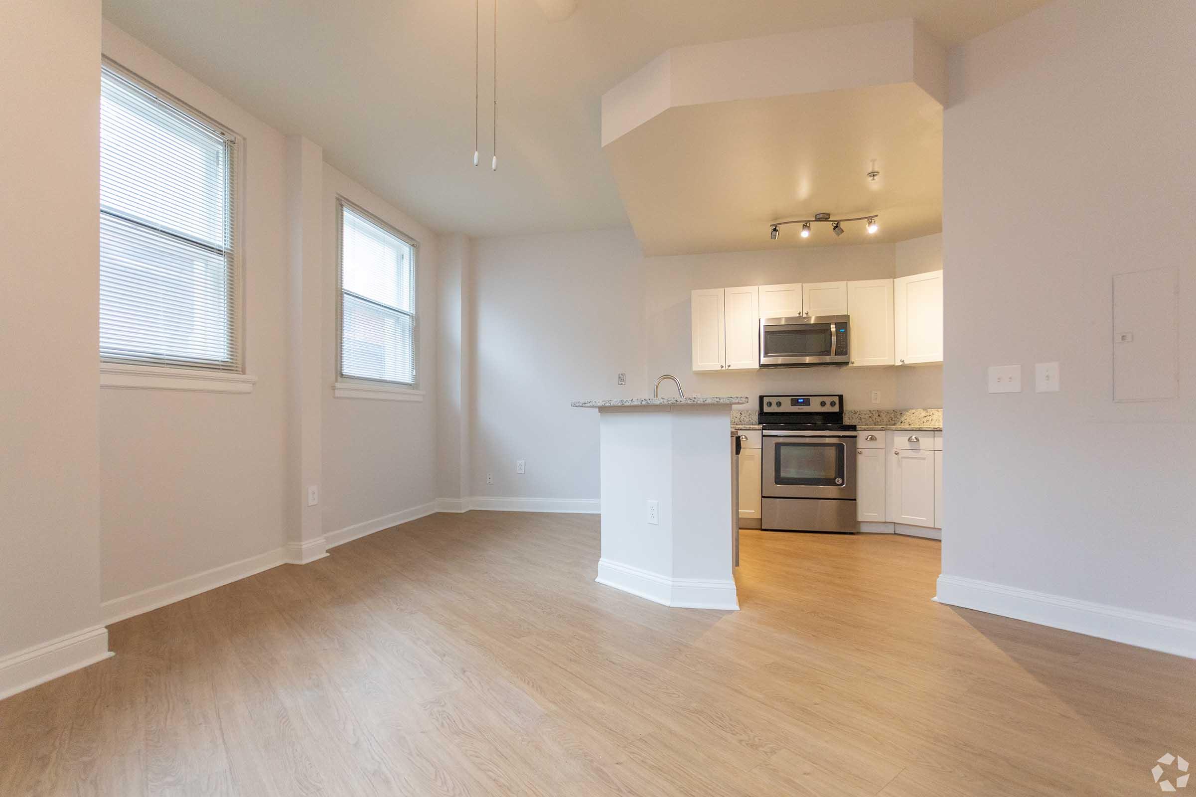 Spacious modern living area featuring light-colored walls, large windows allowing natural light, and a kitchen with stainless steel appliances and a granite countertop. The floor has a warm wood finish, creating a welcoming atmosphere in a bright and open layout.