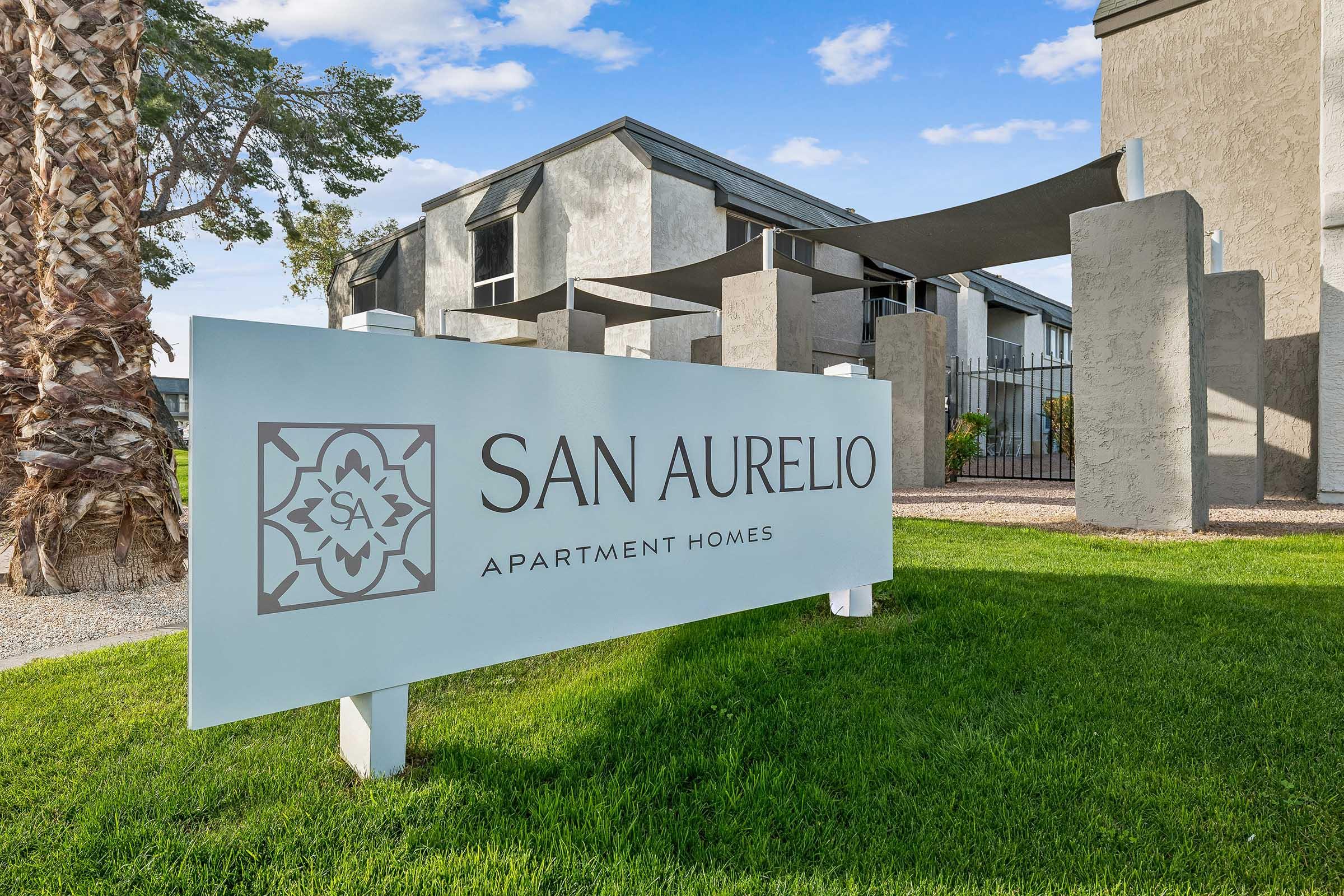 Sign for San Aurelio Apartment Homes, featuring modern design elements with the name prominently displayed. The background includes a landscaped area with palm trees and the building structure partially visible. The scene is set against a bright sky, indicating a welcoming environment.