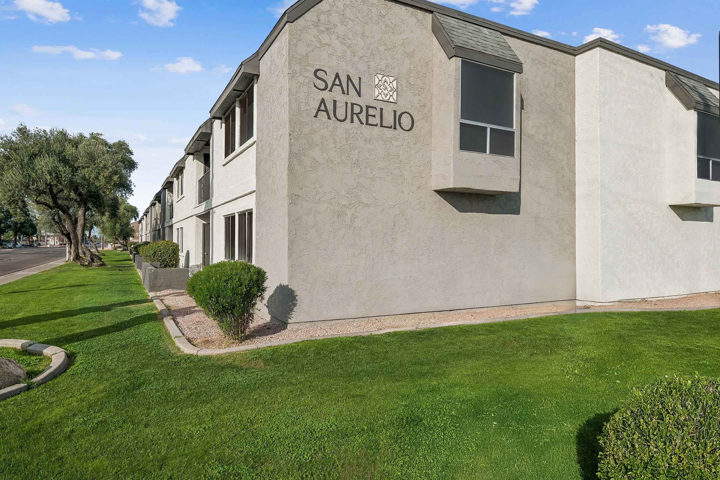 Exterior view of a multi-unit residential building with the name "San Aurelio" prominently displayed on the wall. The building features a modern architectural design, with grassy landscaping, shrubs, and a clear blue sky in the background.