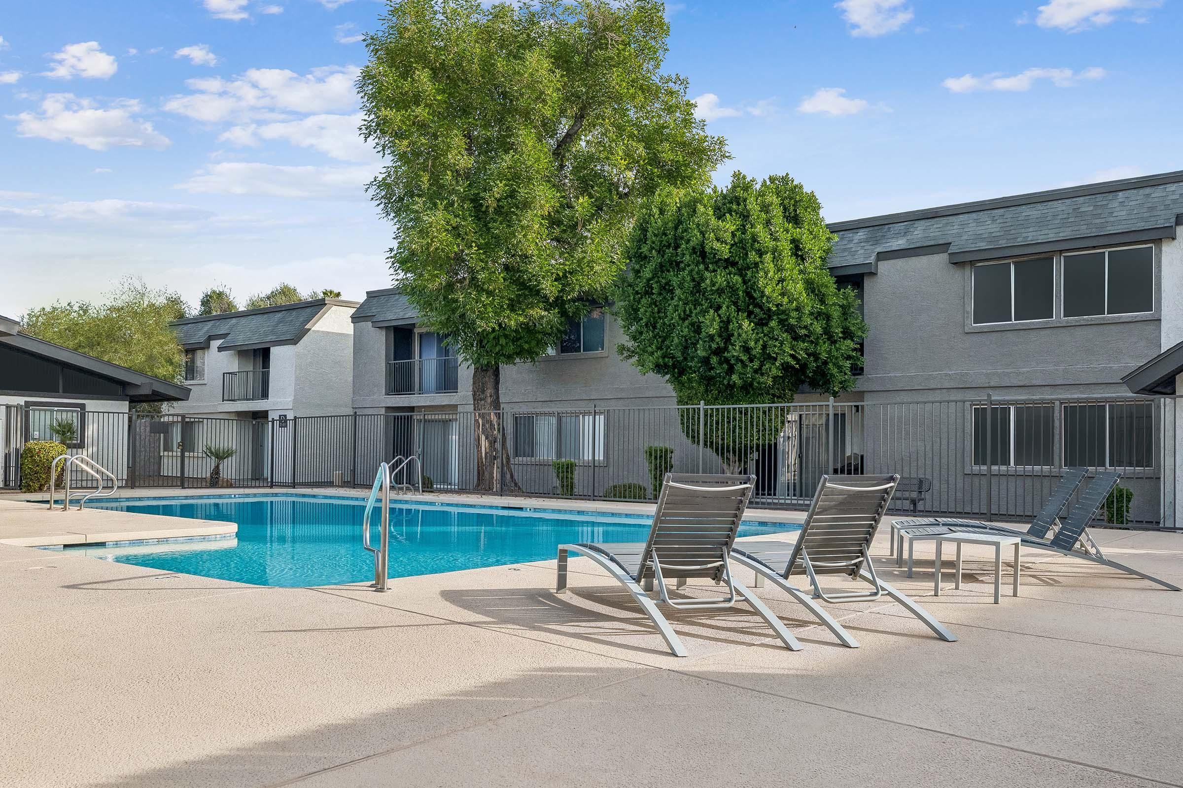 A view of a swimming pool surrounded by lounge chairs, with two large trees nearby. The pool area is enclosed by a fence, and in the background, there are two-story apartment buildings with multiple windows. The sky is clear with a few clouds, creating a serene atmosphere.