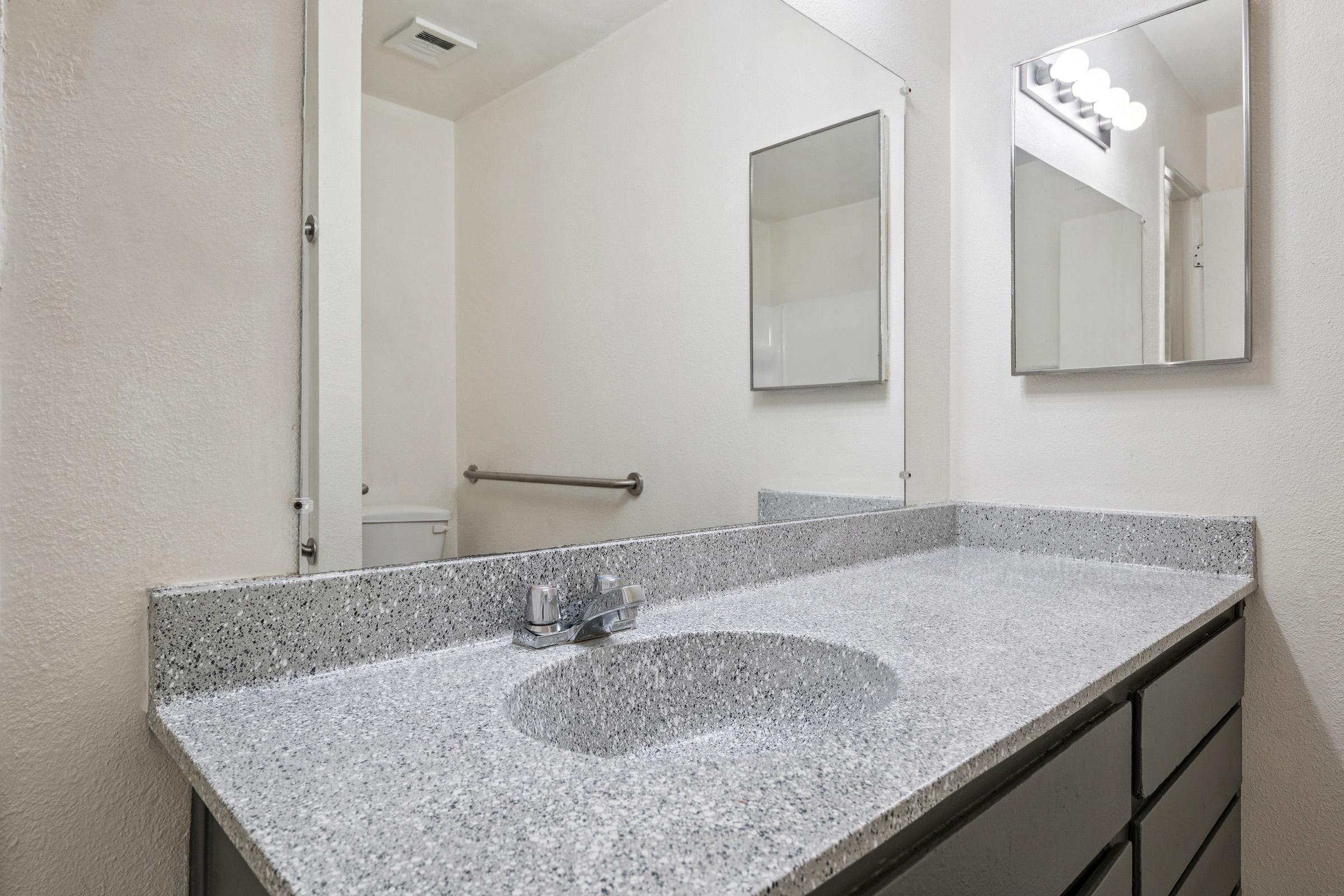 A modern bathroom sink area featuring a gray speckled countertop, a single chrome faucet, and a round sink. There are two mirrors above the sink, and the background includes a white wall and an offering space for a handrail, suggesting a functional and accessible design.