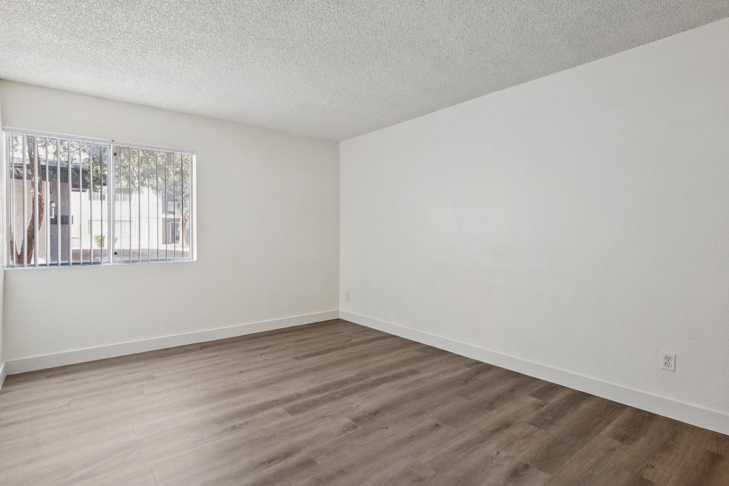 A vacant room with light-colored walls and a window featuring vertical blinds. The floor is covered with light wood laminate, and there are no furnishings or decorations in the space. Natural light is streaming in through the window, creating a bright atmosphere.