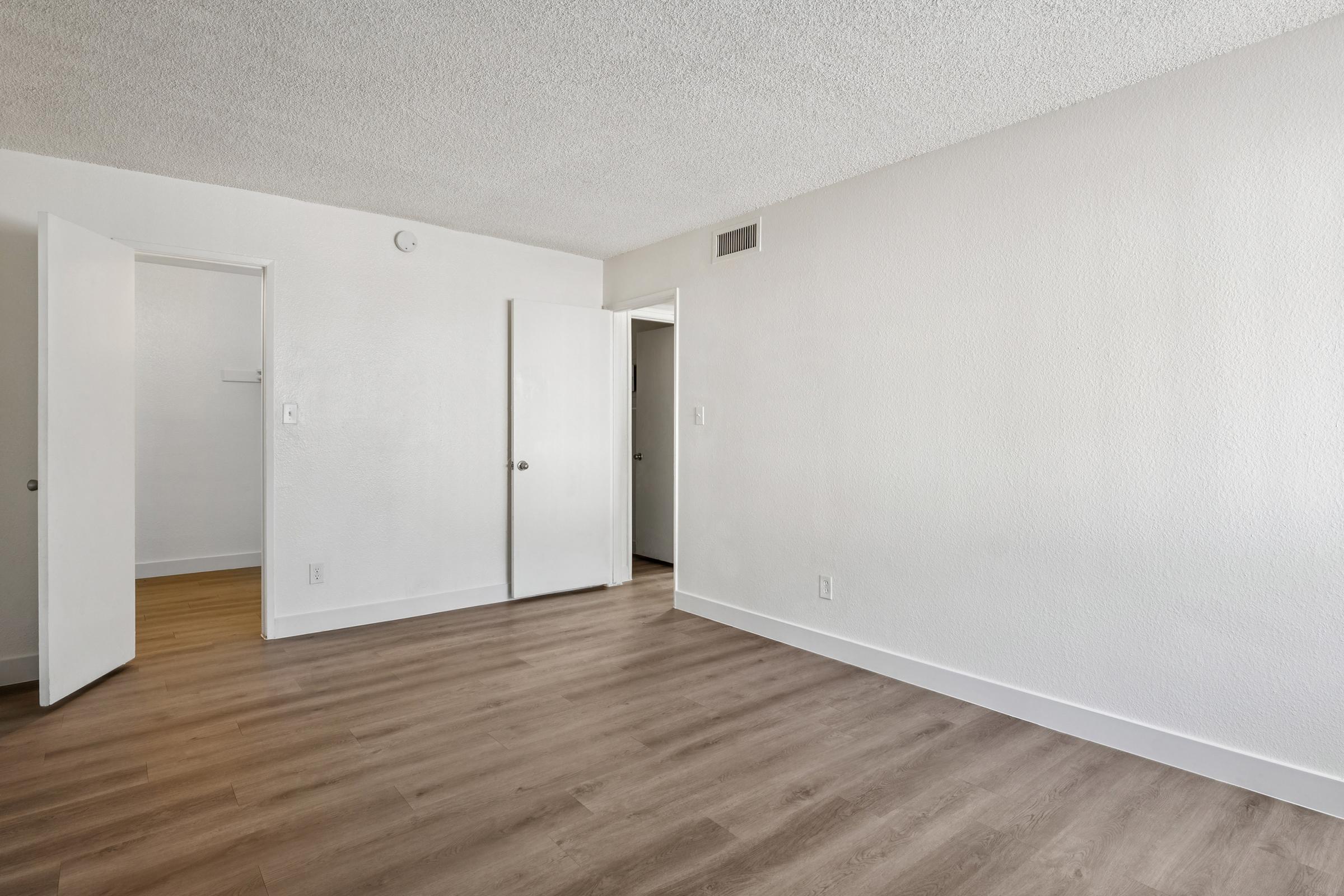 A spacious empty room with light-colored walls and a textured ceiling. There are two open doors leading to adjacent spaces, and the floor is finished with light wood laminate. Natural light fills the room, creating a bright and airy atmosphere.