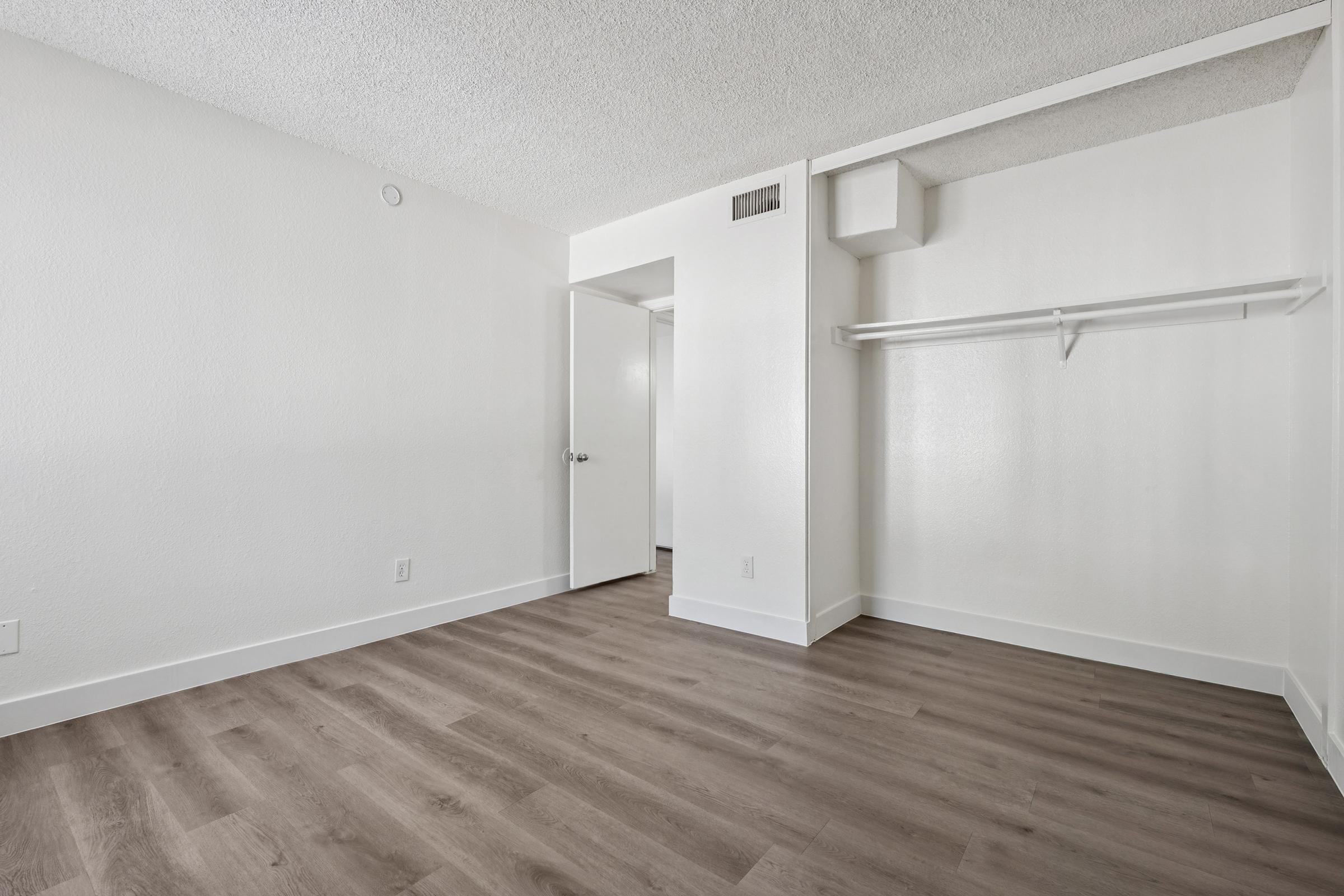 Empty room with light-colored walls and wooden flooring. A closet with a hanging rod is visible on the left, and a closed door is positioned to the right. The room has a simple, minimalistic design and good natural light, creating a spacious feel.