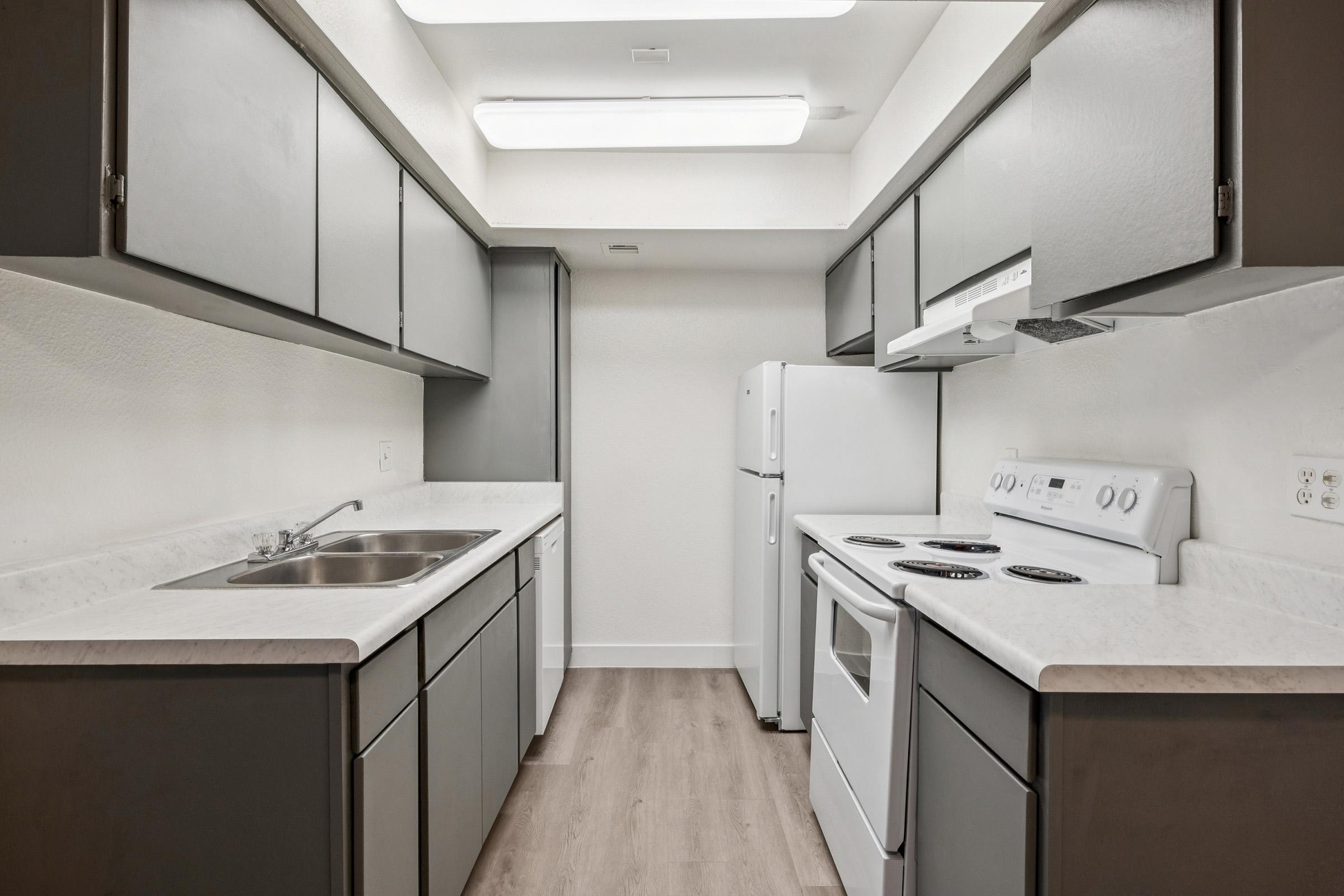 A modern kitchen featuring gray cabinets, white countertops, a double sink, and an electric stove. There is a white refrigerator and an oven underneath the microwave. The walls are painted light, and the flooring is a light wood laminate, giving the space a clean and contemporary look.
