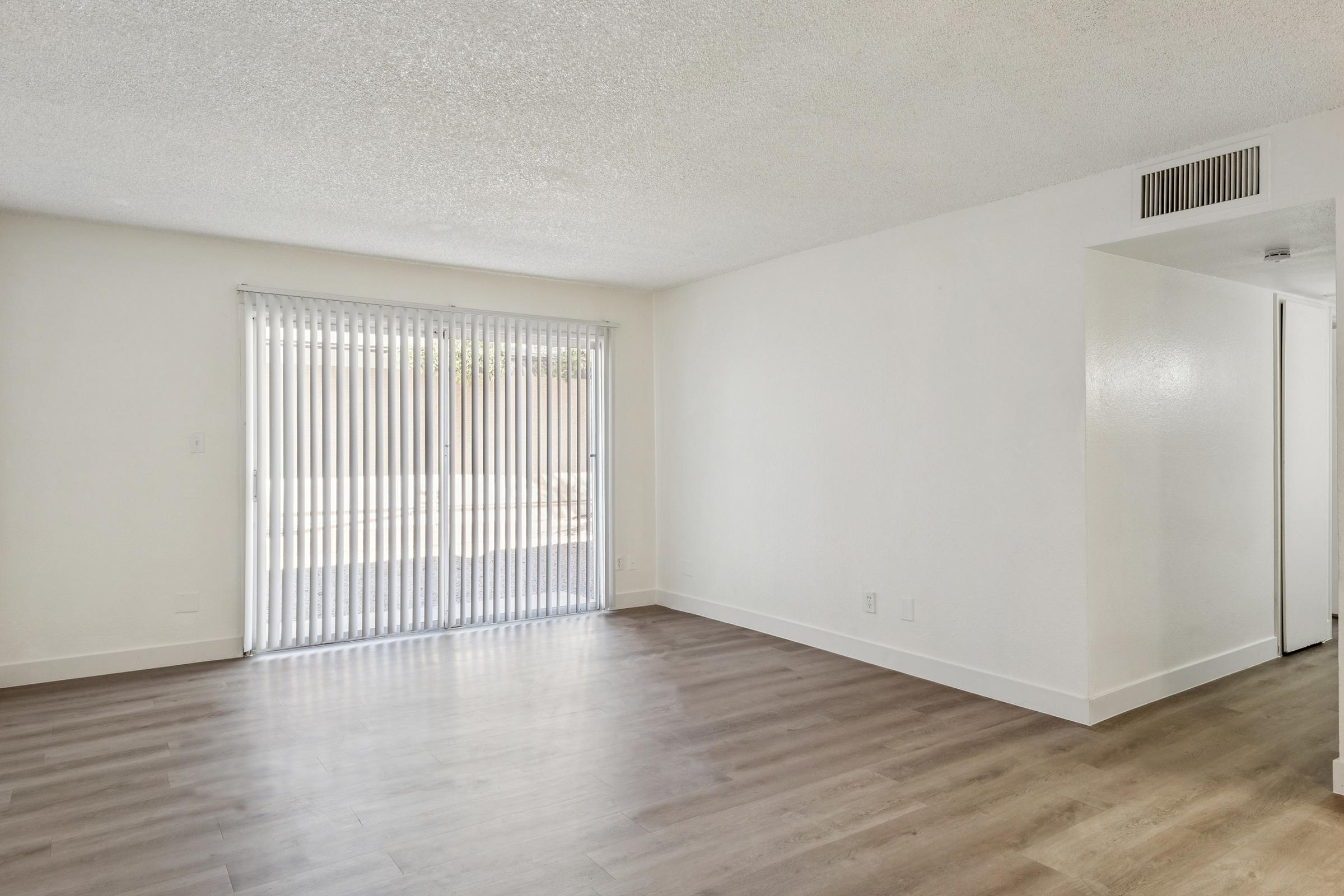 A spacious room with light-colored walls and a textured ceiling, featuring a large sliding glass door with vertical blinds that allows natural light to enter. The floor is covered in light-colored wood laminate. A door on the right leads to another area, adding to the room's open feel.