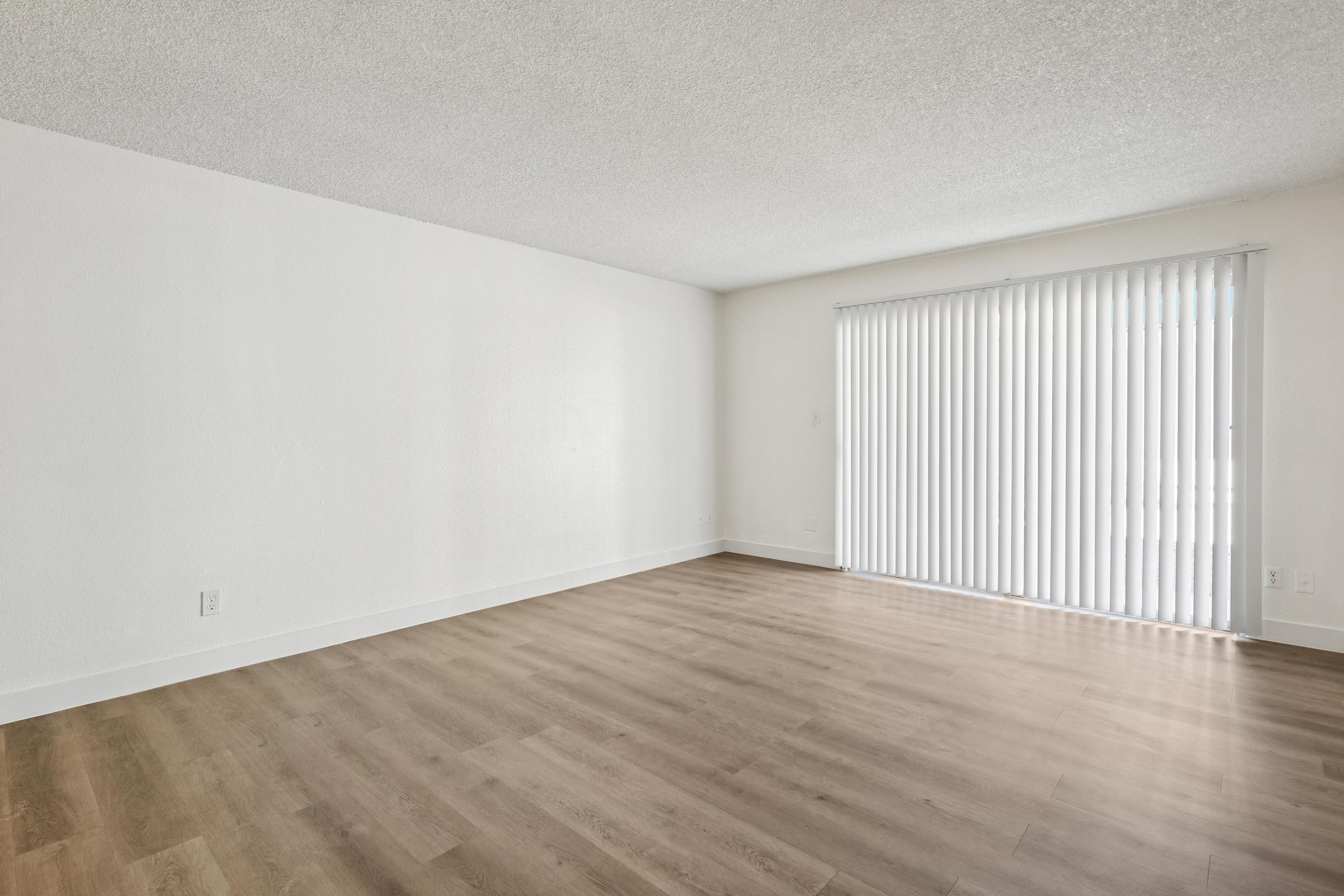 Empty room with light-colored walls and laminate flooring. A large window with vertical blinds allows natural light to fill the space, creating a bright and airy atmosphere. The room is devoid of furniture, showcasing the open and spacious layout.