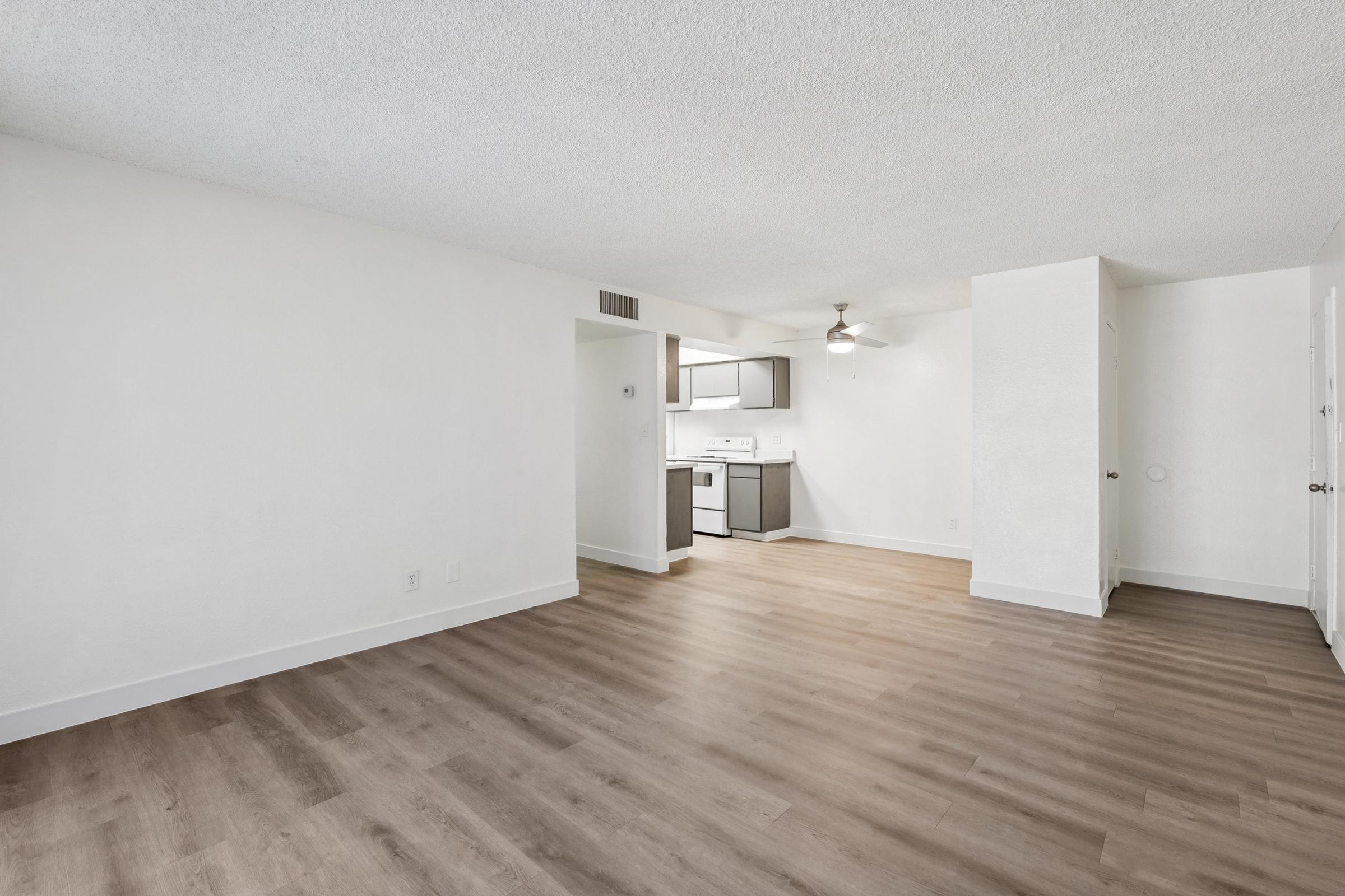 Spacious living area with light-colored walls and a textured ceiling. The room features laminate flooring and a ceiling fan. A doorway leads to a kitchen area with modern appliances and cabinetry visible in the background. Natural light brightens the space, creating an inviting atmosphere.