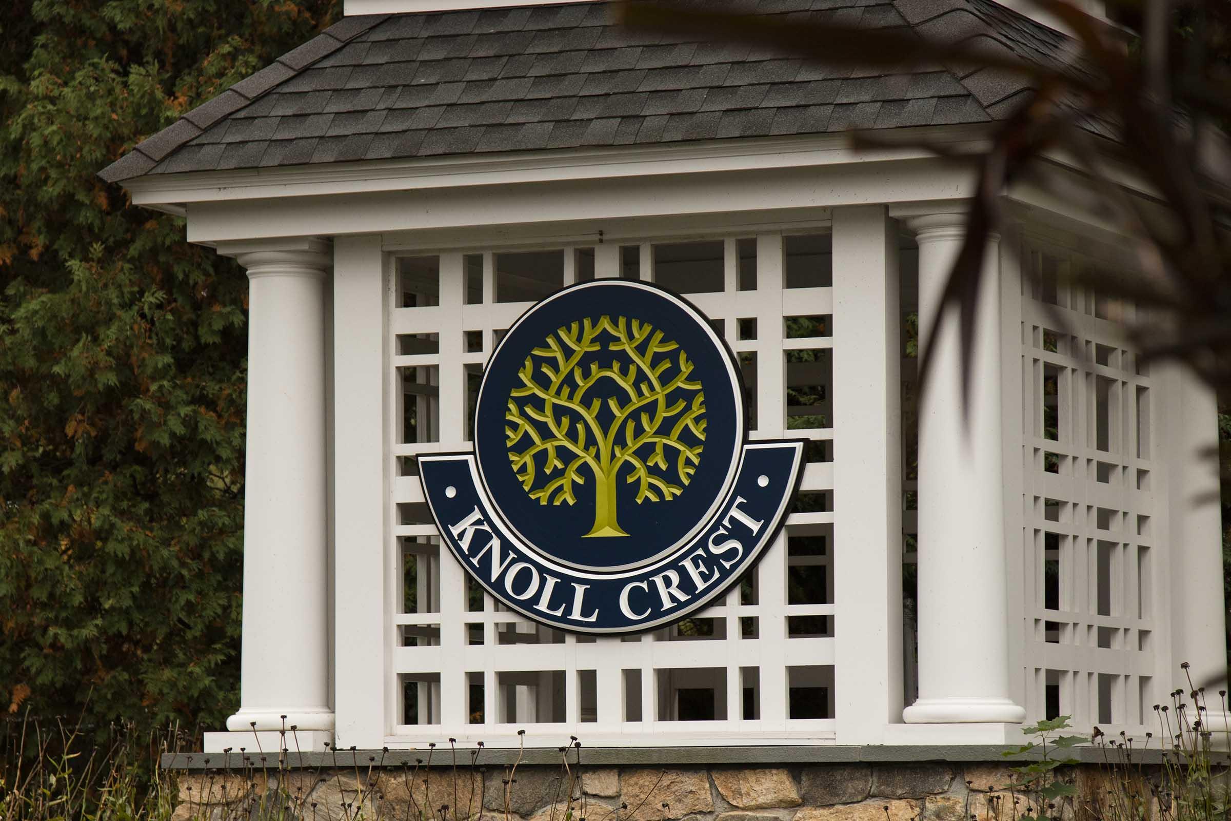 A decorative sign displaying "Knoll Crest" featuring a circular logo with a tree design. The sign is mounted on a white structure with a lattice-style background, surrounded by greenery.