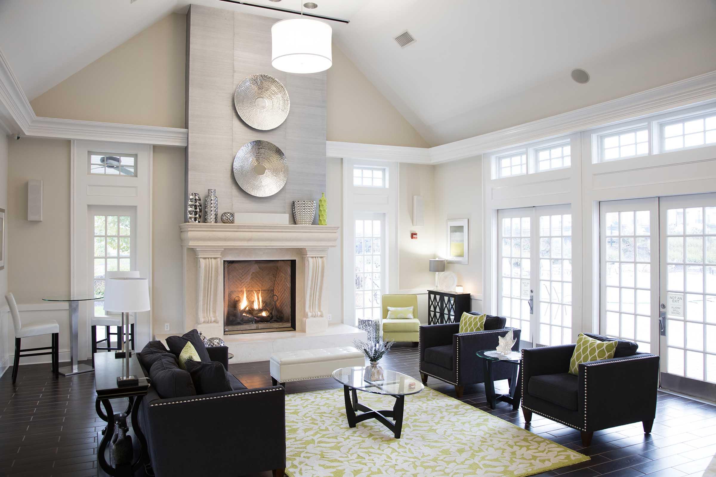 A spacious living room with a cozy fireplace as the focal point. The room features modern furniture, including black sofas and armchairs, a light-colored coffee table, and decorative pillows in green and white. Large windows allow natural light to fill the space, complemented by a stylish area rug.