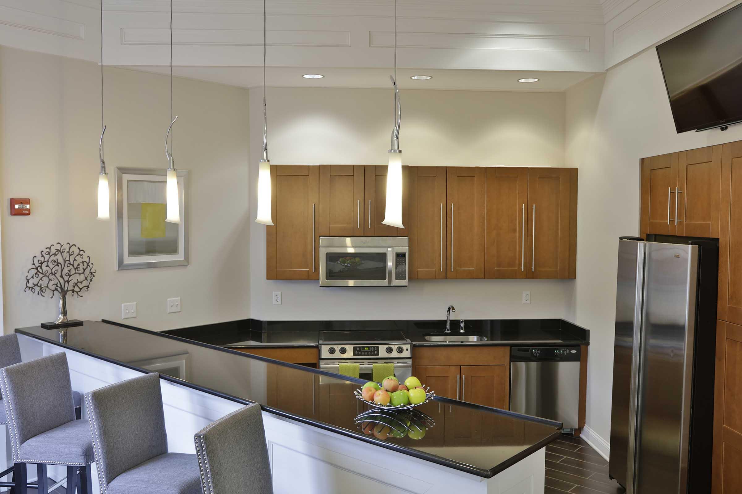 A modern kitchen featuring wooden cabinetry, stainless steel appliances, and a black countertop. The space includes a sleek island with bar stools, decorative fruit bowl, and pendant lighting, creating a bright and inviting atmosphere. A television is mounted on the wall, enhancing the room's functionality.