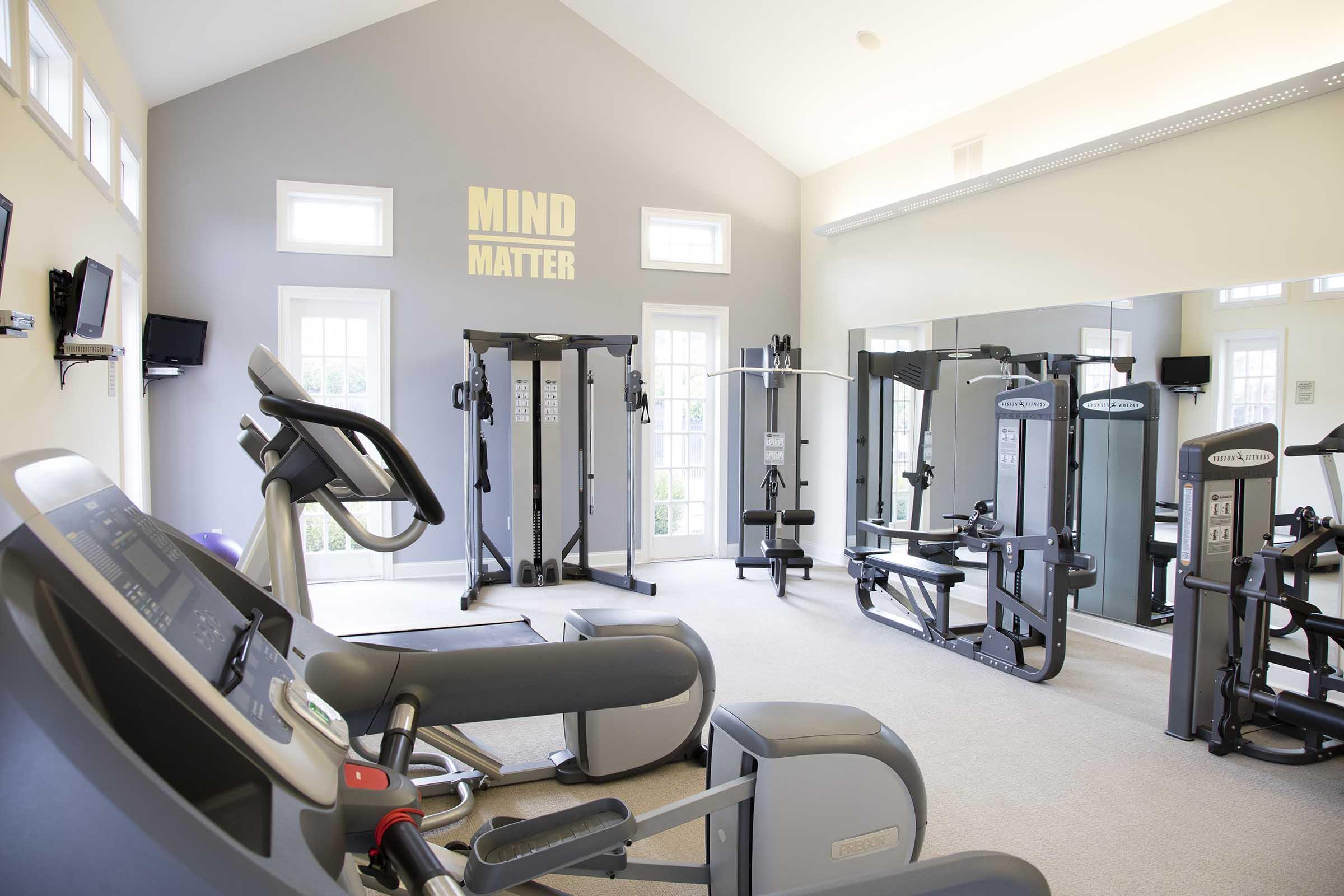A modern fitness room featuring various exercise equipment, including a stationary bike, weight machines, and mirrors. The walls are painted light gray with the words "MIND" and "MATTER" prominently displayed. Large windows allow natural light to illuminate the space.