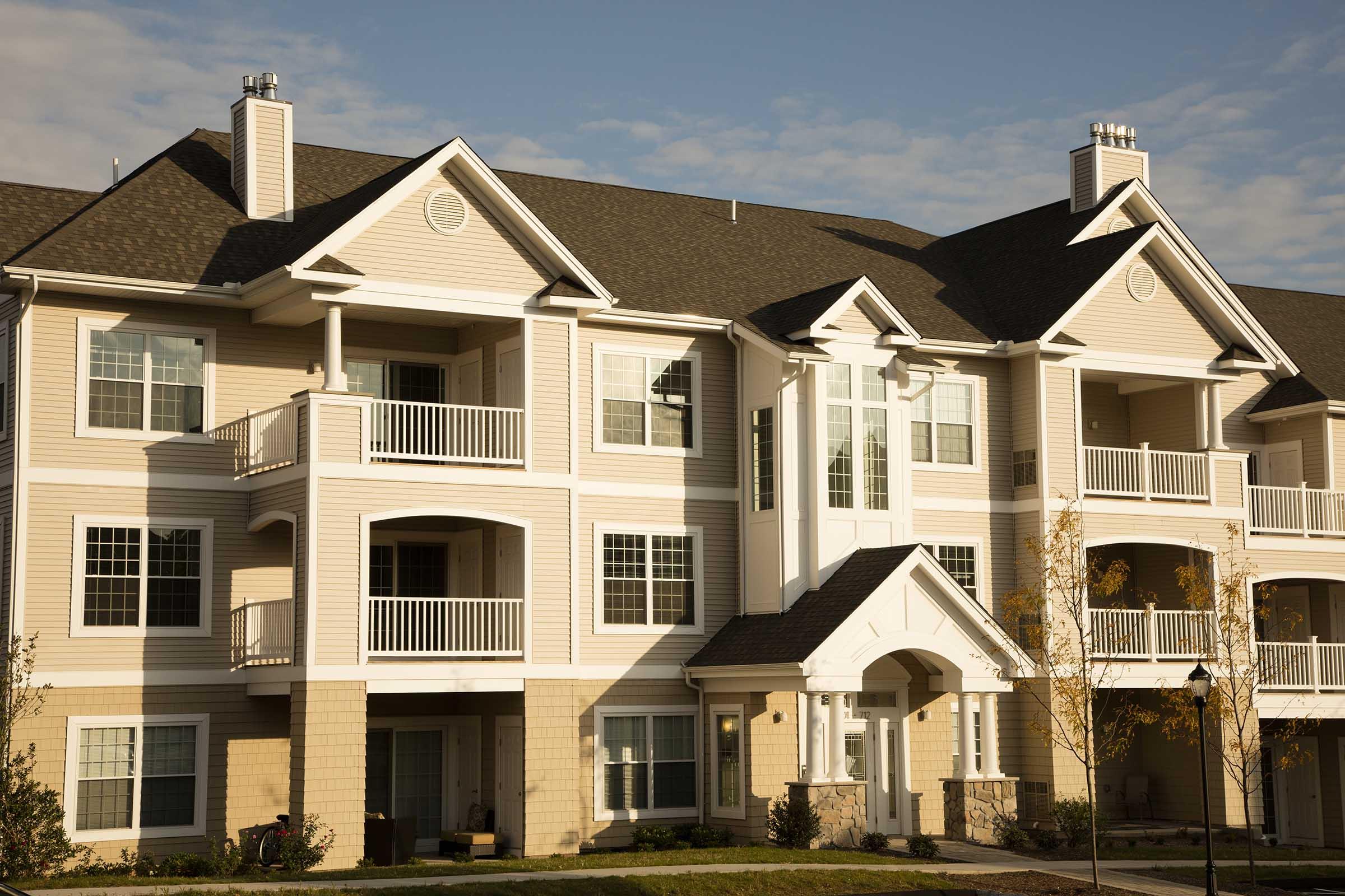 A modern multi-unit residential building featuring beige siding, white railings on balconies, and a three-story design. The architecture includes decorative elements like pillars and a prominent entrance, set against a clear blue sky. Lush green landscaping enhances the exterior.