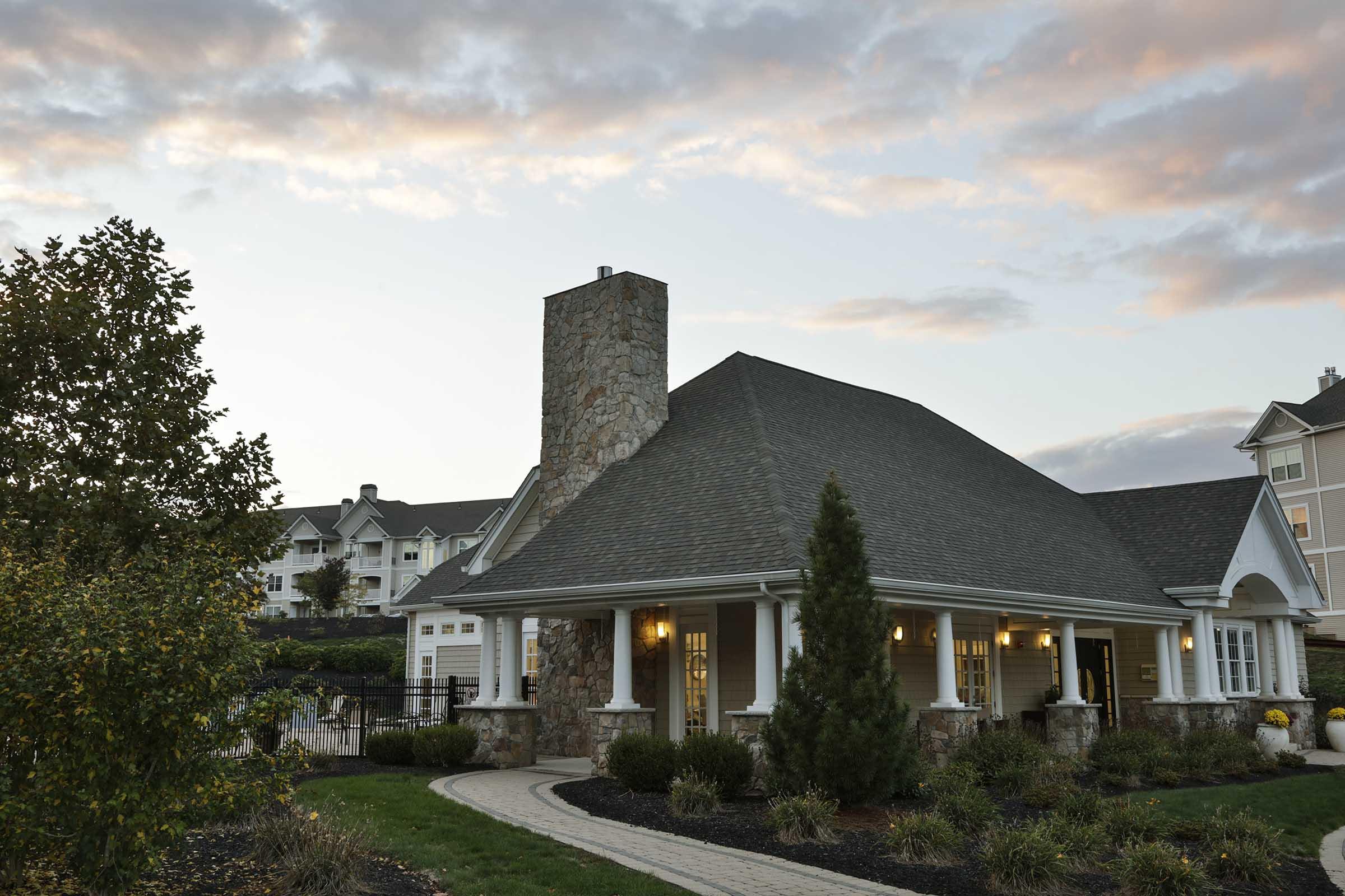 A cozy stone-fronted building with a sloped roof and a chimney, surrounded by landscaped greenery and colorful flowers. The sky is filled with soft clouds, hinting at twilight. In the background, there are other residential buildings, creating a serene community atmosphere.