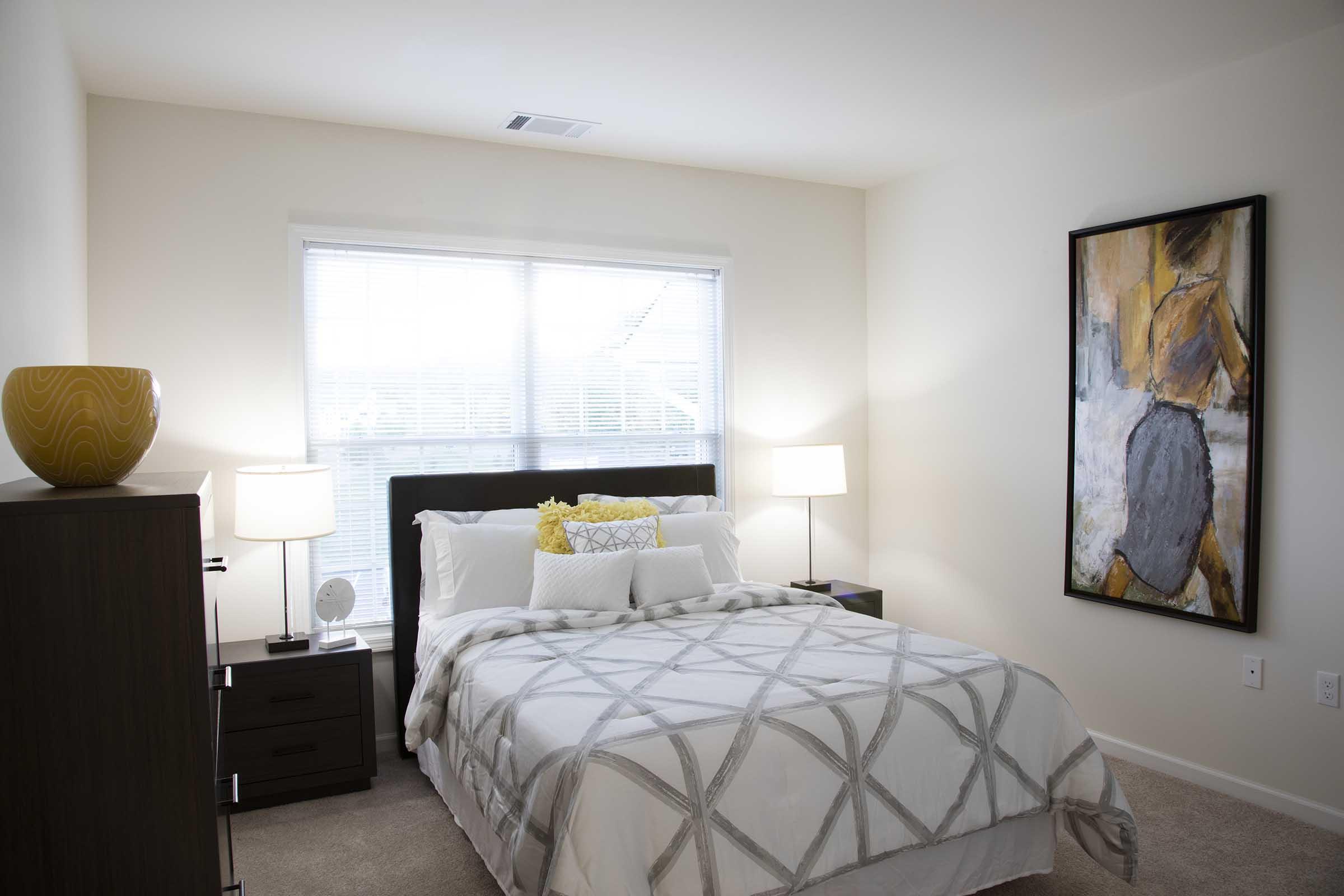 A well-decorated bedroom featuring a queen-sized bed with a light grey and white patterned comforter, a yellow accent pillow, two bedside lamps on dark wood nightstands, a decorative bowl on the dresser, and a large abstract painting on the wall. Natural light streams through a window, creating a cozy atmosphere.