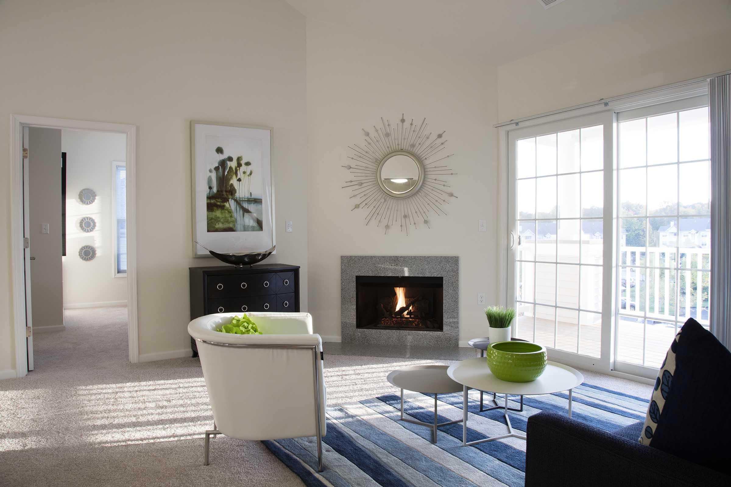 A modern living room featuring a cozy fireplace, a large mirror, and stylish furniture. There is a white chair and a coffee table with green decor. The room has large windows allowing natural light, with a soft carpet and decorative wall art creating an inviting atmosphere.