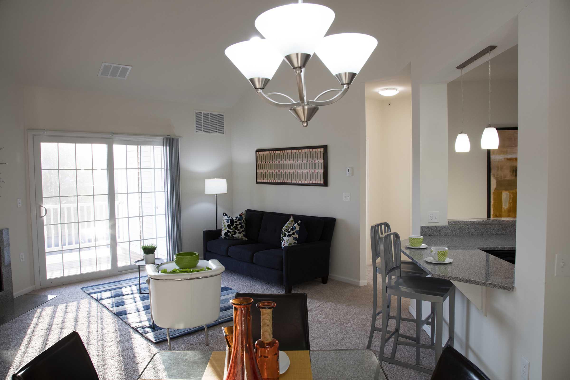 A modern living room and dining area featuring a dark sofa, a glass dining table with decorative items, and a breakfast bar with stools. Natural light pours in through sliding glass doors, illuminating the space. Warm tones and contemporary decor create a cozy atmosphere.