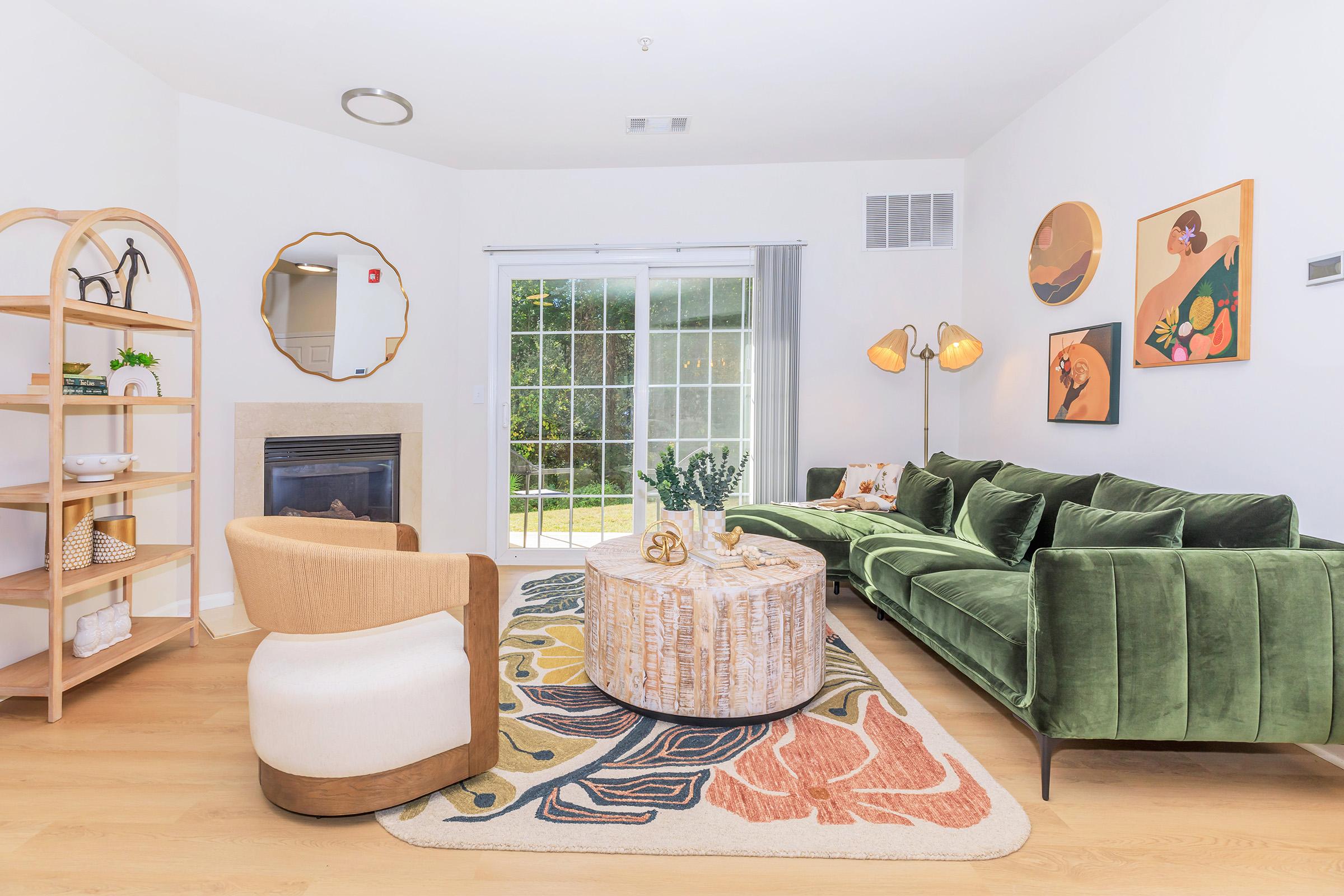 A modern living room featuring a green velvet sofa, a round coffee table with a textured design, and a beige accent chair. The decor includes a stylish mirror, wall art with floral themes, and a decorative bookshelf. Natural light streams in through large sliding glass doors leading to an outdoor space.