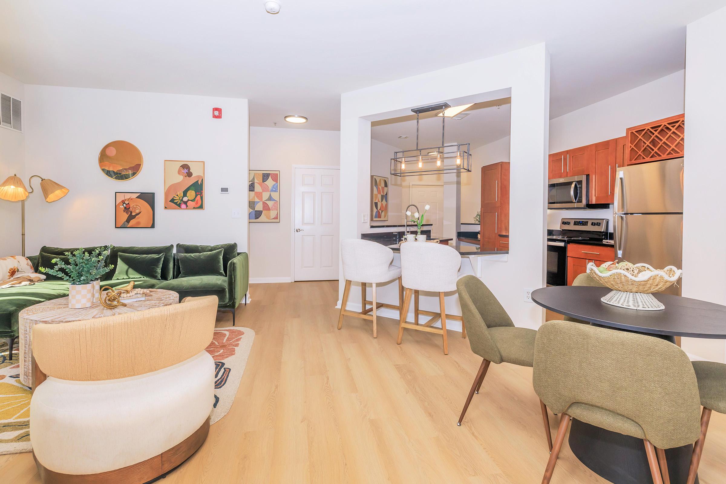A modern living space featuring a green velvet sofa, stylish armchairs, and a round dining table. The open layout includes a kitchen with stainless steel appliances and wooden cabinets. Decorative wall art and a cozy rug enhance the inviting atmosphere of the room. Bright and airy with natural light.