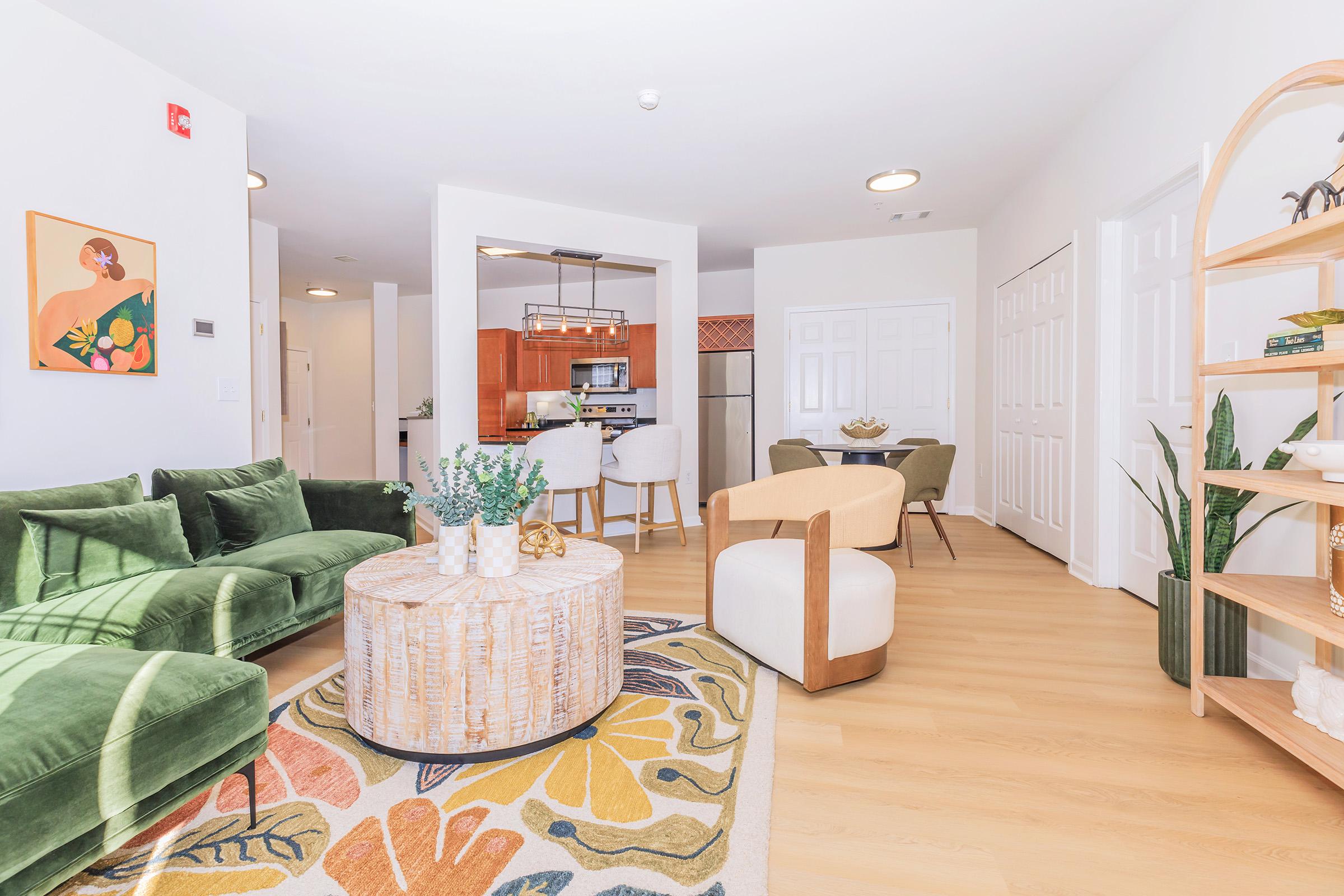 Modern living room featuring a plush green sofa, a round coffee table with floral design, and a stylish accent chair. The space includes a dining area with chairs, a kitchen in the background, and indoor plants for a fresh touch. Bright and airy atmosphere with light colors enhancing the decor.