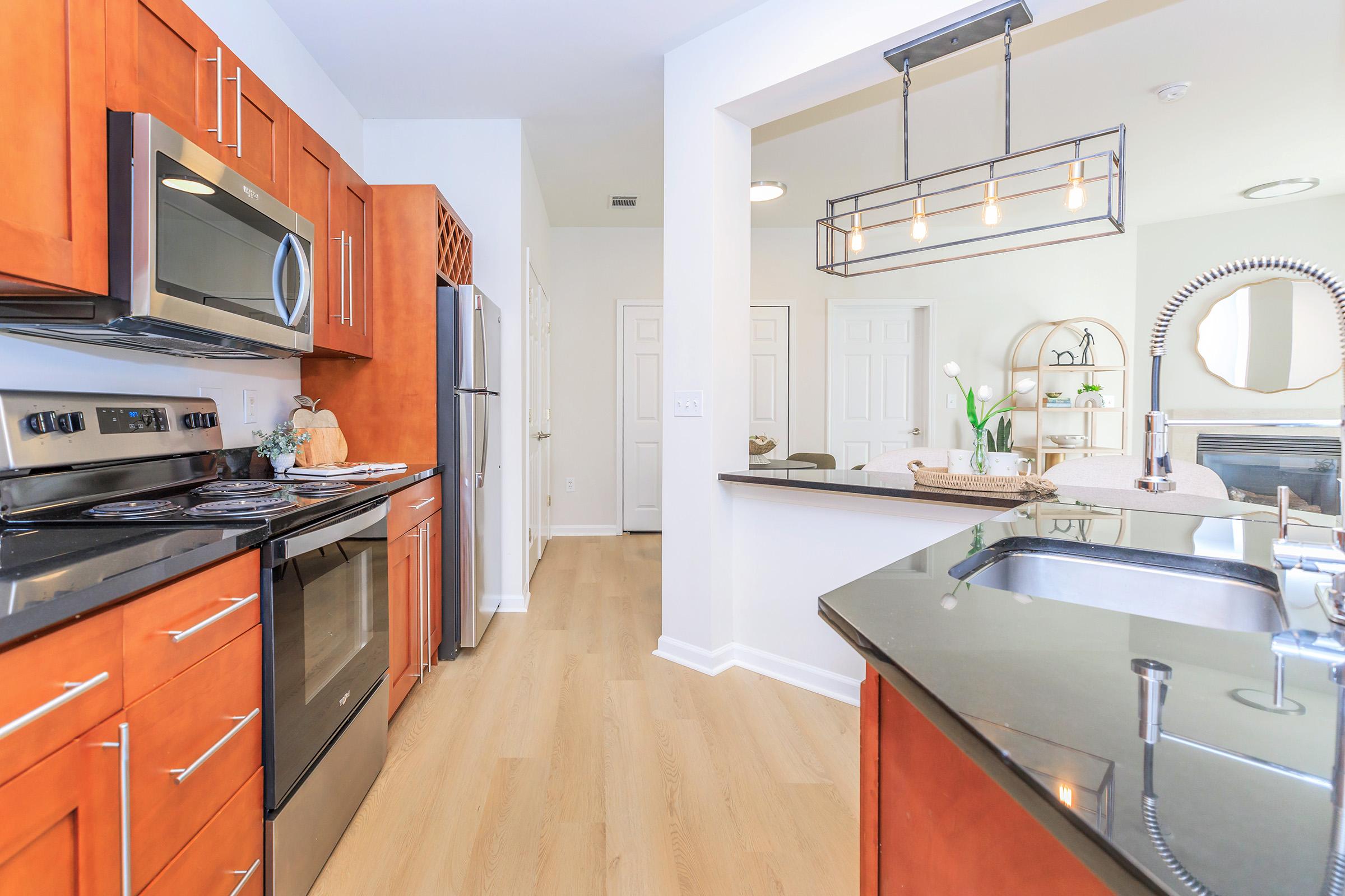 Modern kitchen with wooden cabinets, stainless steel appliances, and a black countertop. A sink is visible, along with a kitchen island. Bright lighting and a doorway lead to an adjacent room, creating an open and spacious feel. Fresh flowers are placed decoratively on the counter.