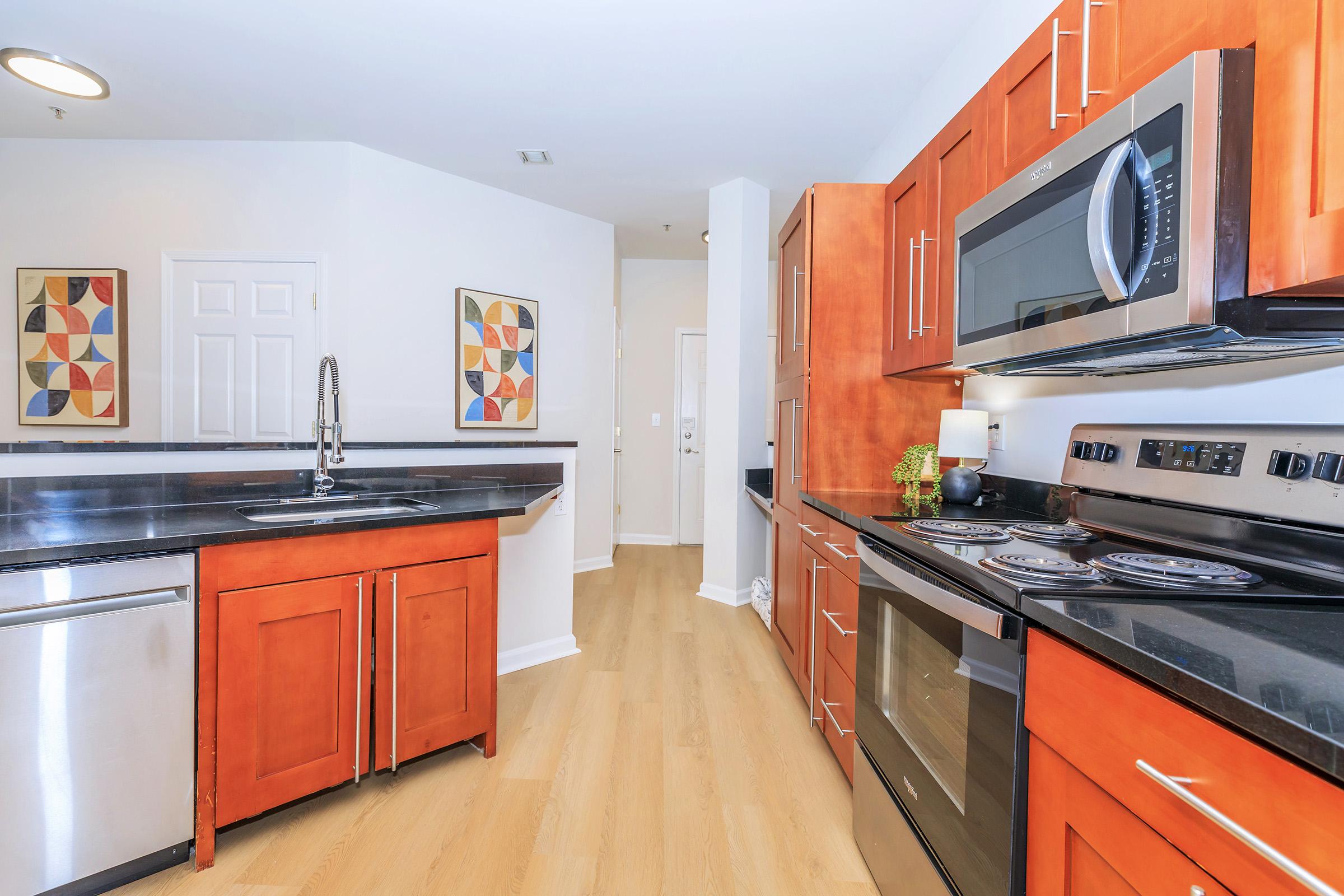 Modern kitchen featuring wooden cabinets with a warm orange finish, stainless steel appliances, and a dark granite countertop. The design includes an island and a window, creating an open and inviting space. The walls are decorated with geometric artwork for a contemporary touch.