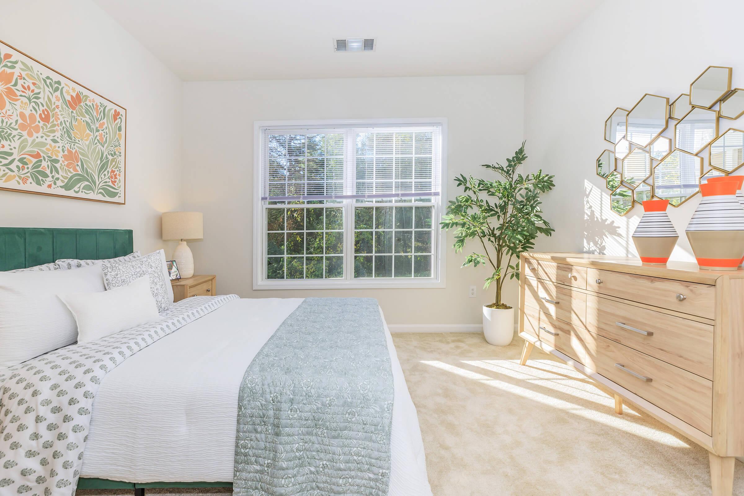 A bright and inviting bedroom featuring a large window with natural light, light-colored walls, a green headboard, a cozy bed with decorative pillows, and a soft throw blanket. There is a wooden dresser with stylish decor, including geometric mirrors and potted plants, creating a warm and relaxing atmosphere.