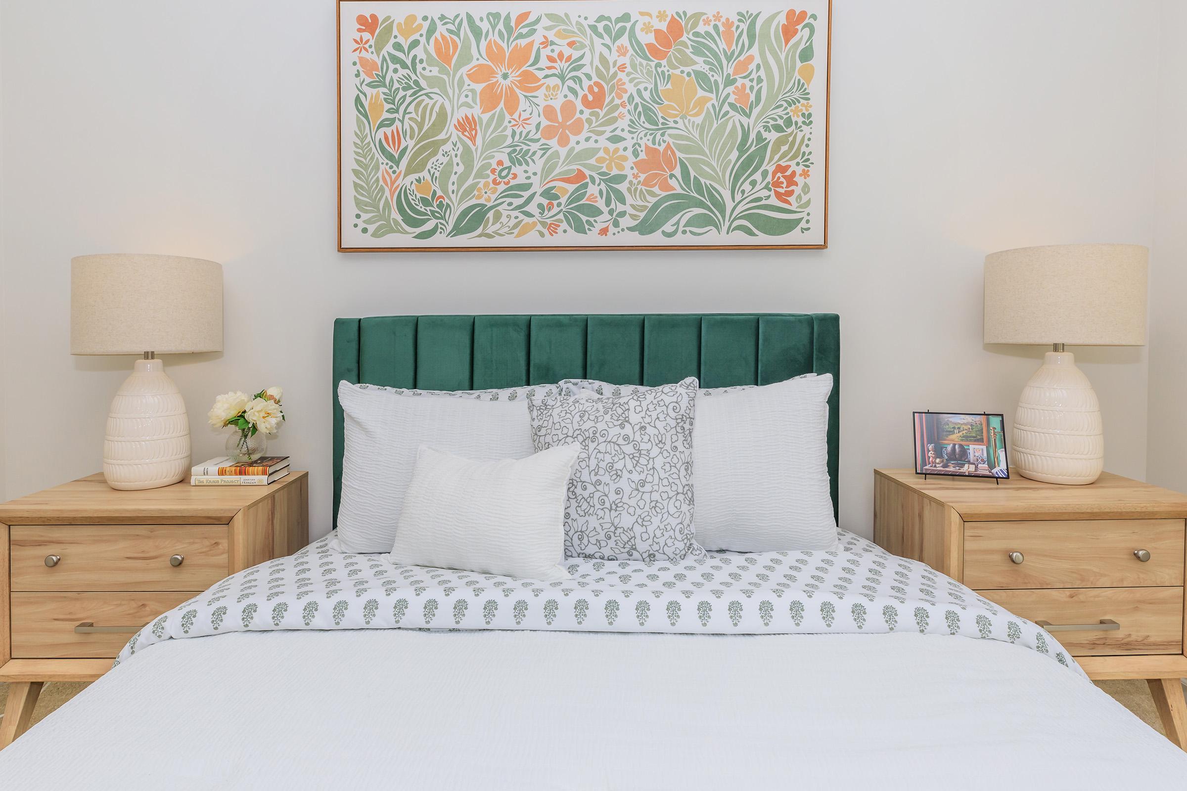 A cozy bedroom featuring a green tufted headboard, neatly arranged pillows, and a light-colored bedding set. On either side of the bed are wooden nightstands with lamps and decorative items, including a framed photo and a small bouquet of flowers. A floral wall art piece adds a vibrant touch above the headboard.