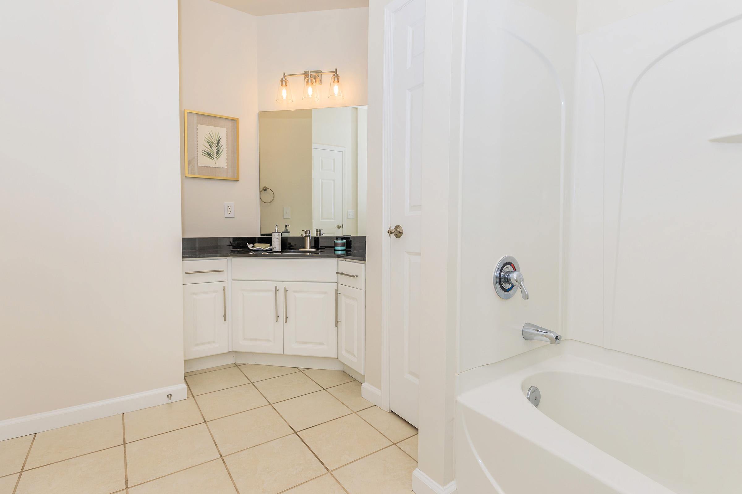A modern bathroom with a cream-colored tiled floor. It features a large mirror above a double-sink vanity with stylish fixtures and a framed botanical print on the wall. To the side, a white bathtub is partially visible, creating a clean and serene aesthetic.