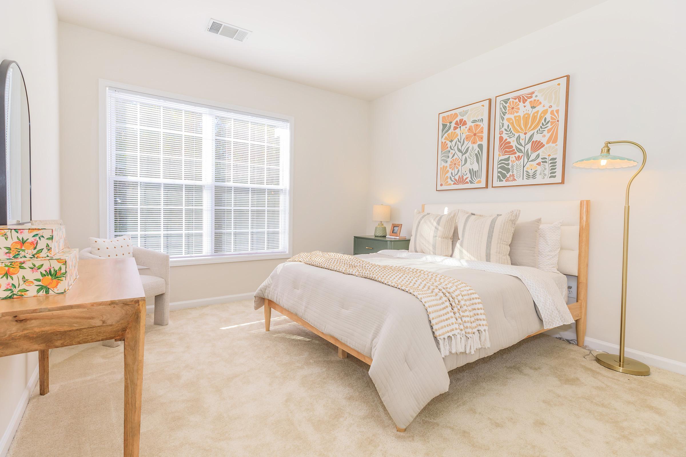 A bright and airy bedroom featuring a large window with white blinds, a neatly made bed with light-colored linens and decorative pillows, a wooden nightstand, a stylish floor lamp, and two framed floral prints on the wall. A small chair and a wooden table with decorative items complete the cozy atmosphere.