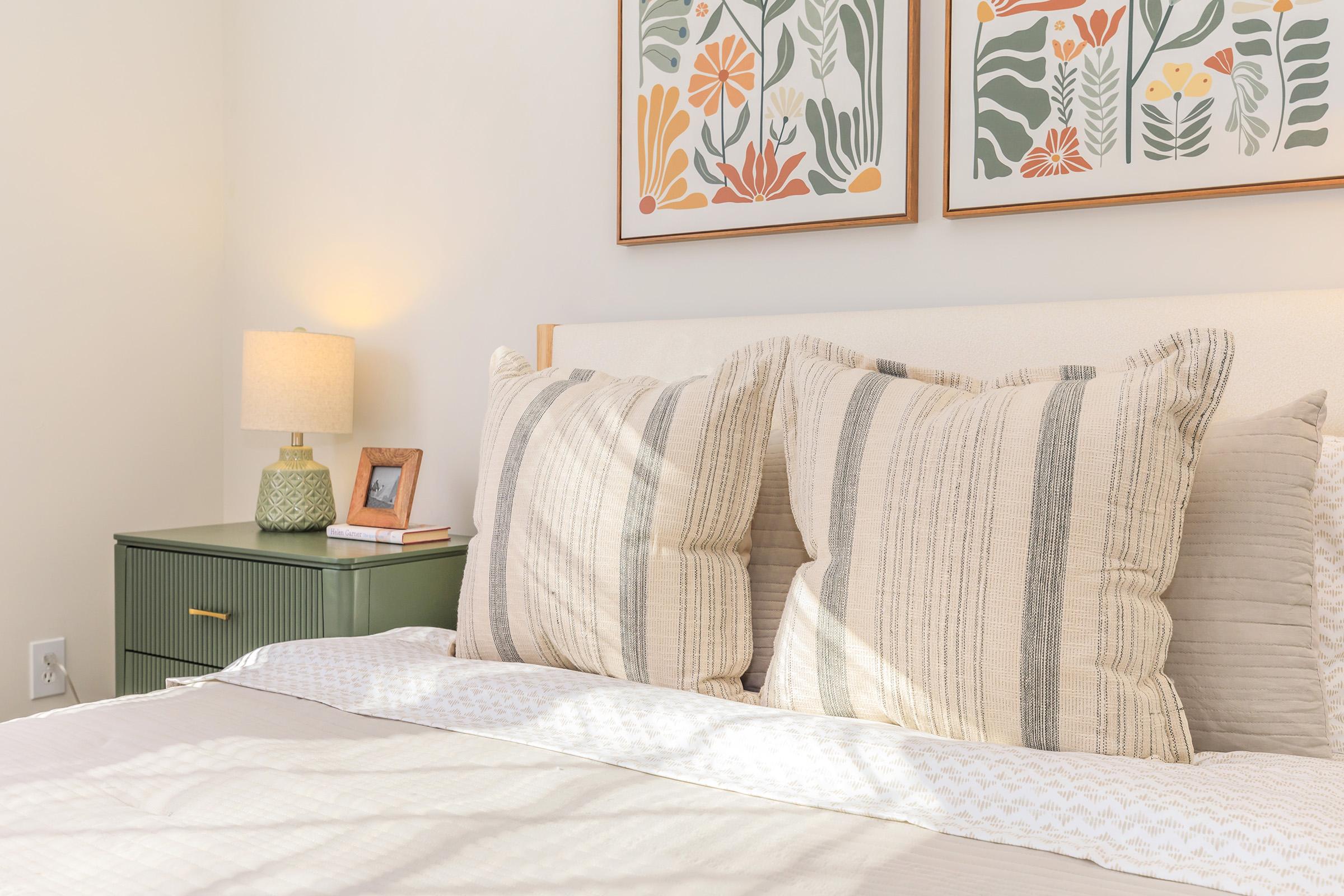 A cozy bedroom featuring a bed with large, textured pillows, a soft quilt, and two floral-patterned art prints on the wall. A small nightstand with a green lamp and a picture frame is beside the bed, creating a warm and inviting atmosphere. Natural light fills the space, enhancing the calming ambiance.