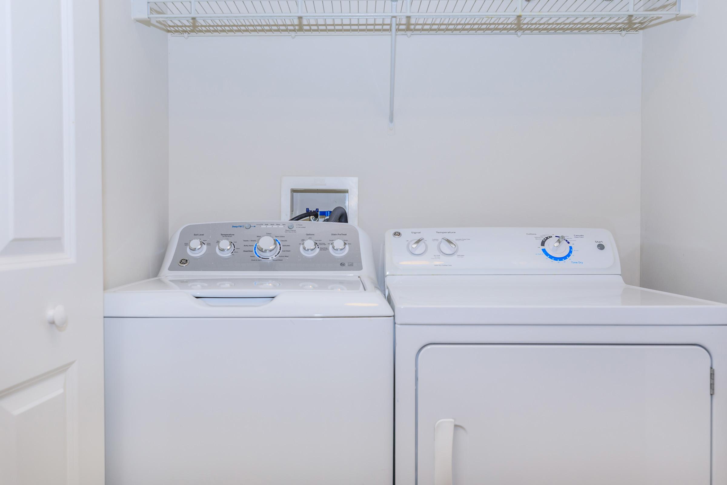 A laundry room featuring a washing machine and a dryer side by side. The machines are white and modern, with control panels that include dials and buttons. There is a wire shelf above the appliances, and a white door partially visible on the left side of the image. The walls are painted white, creating a clean and bright environment.