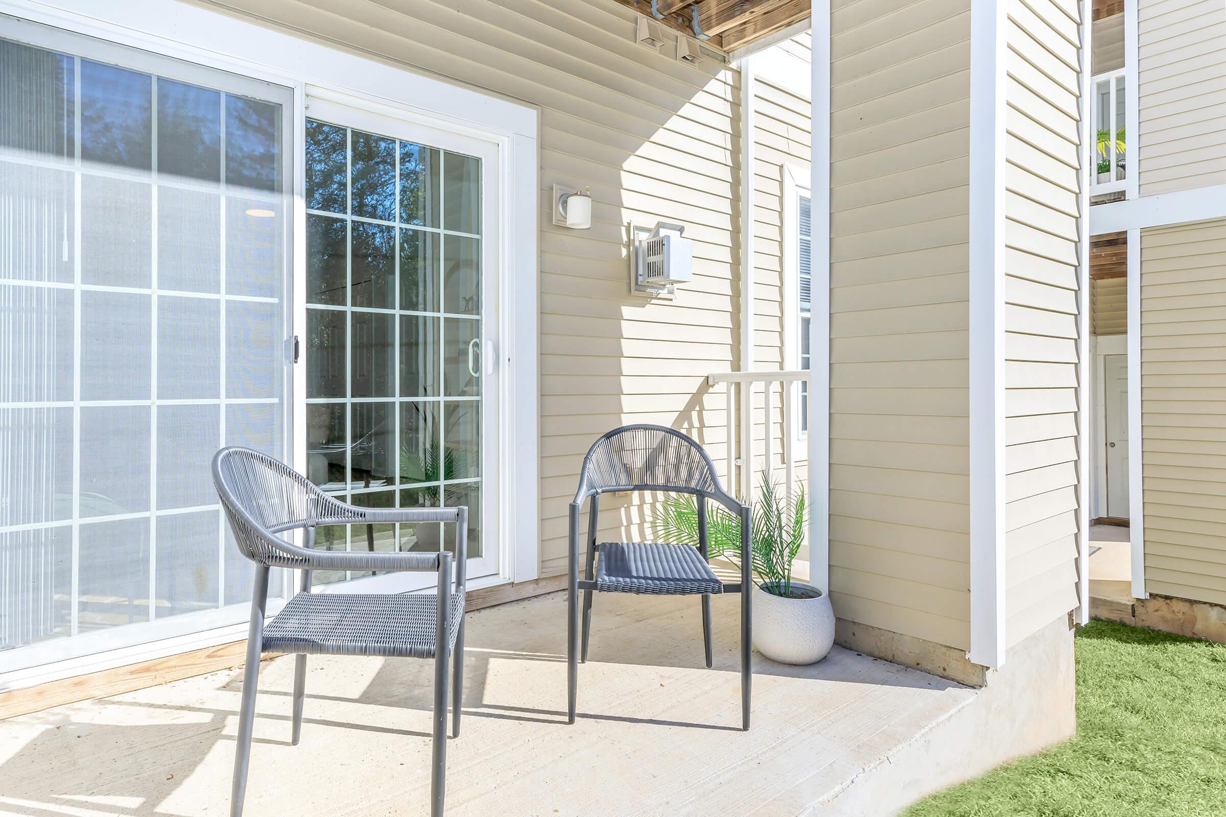 A small outdoor patio area featuring two black chairs beside a modern sliding glass door. The patio is bordered by beige siding and has a potted plant in a white container. The space is well-lit with natural sunlight and includes a grassy area in the background.