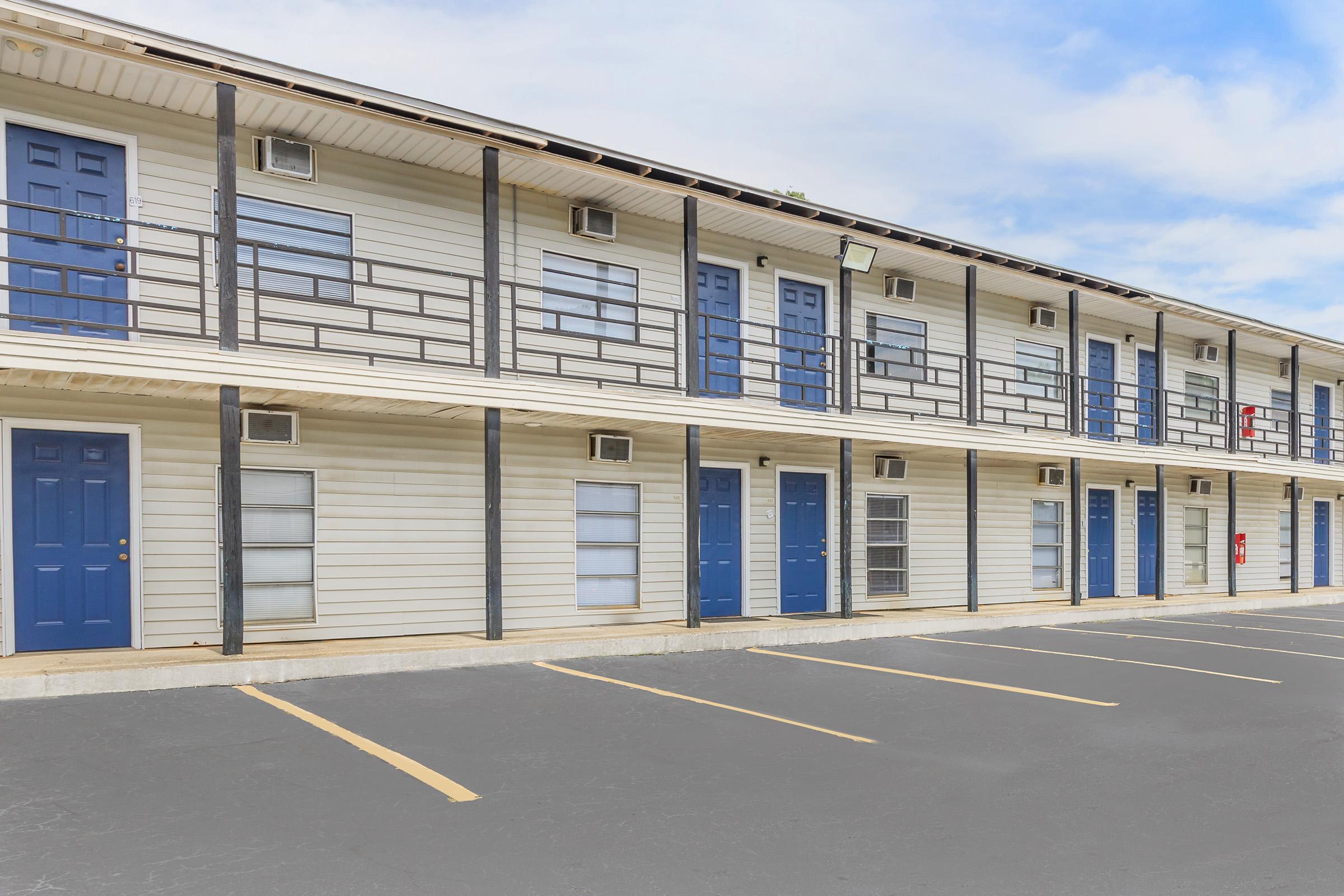Exterior view of a two-story motel with multiple rooms, each featuring blue doors and white siding. The building has a covered walkway with metal railings, and there are several parking spaces in front. The sky is partly cloudy.