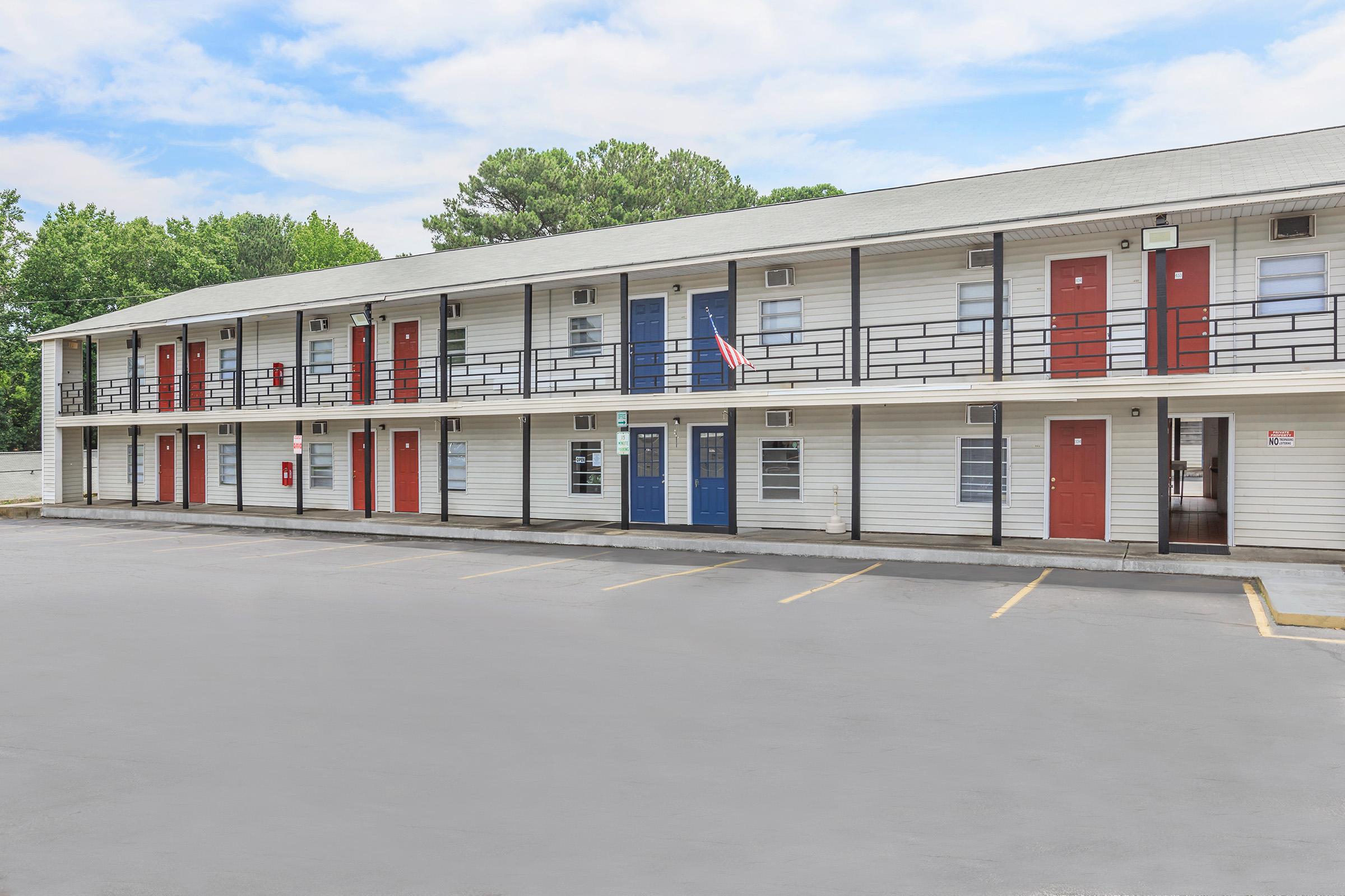 A low-rise motel featuring multiple rooms with white siding and a flat roof. Each room has a door painted in red or blue. There are two American flags displayed, and parking spaces are visible in front of the building. The sky is partly cloudy, and greenery can be seen in the background.