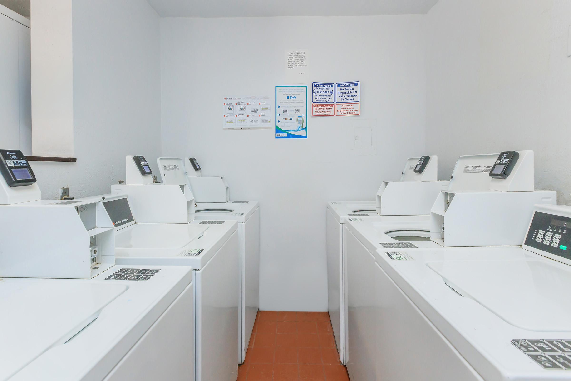 A clean laundry room featuring several white front-loading washing machines arranged in a row. The machines have digital displays and control panels. Light-colored walls surround the space, and there are signs on the wall with instructions and information. The floor is tiled in terracotta.