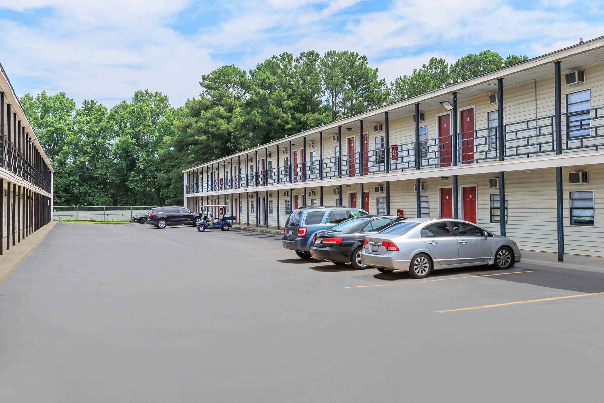 A view of a two-story motel featuring exterior corridors, with several parked cars in a paved lot. The building has red doors and a backdrop of lush green trees under a partly cloudy sky.
