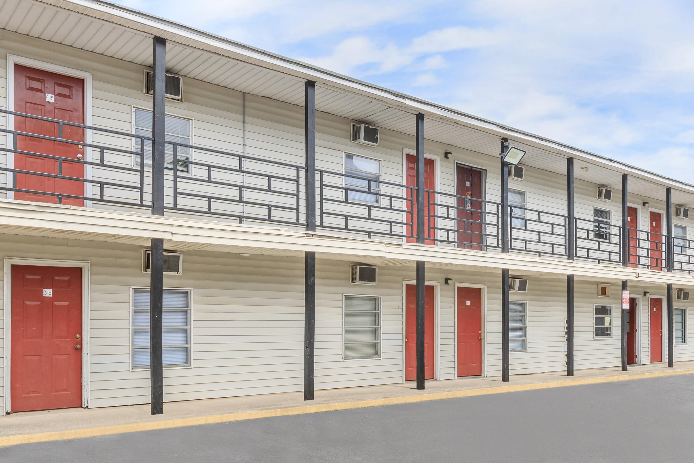 Exterior view of a two-story motel building featuring multiple rooms with red doors and black railings. The facade is light-colored siding with windows visible beside each door. Clear sky in the background.