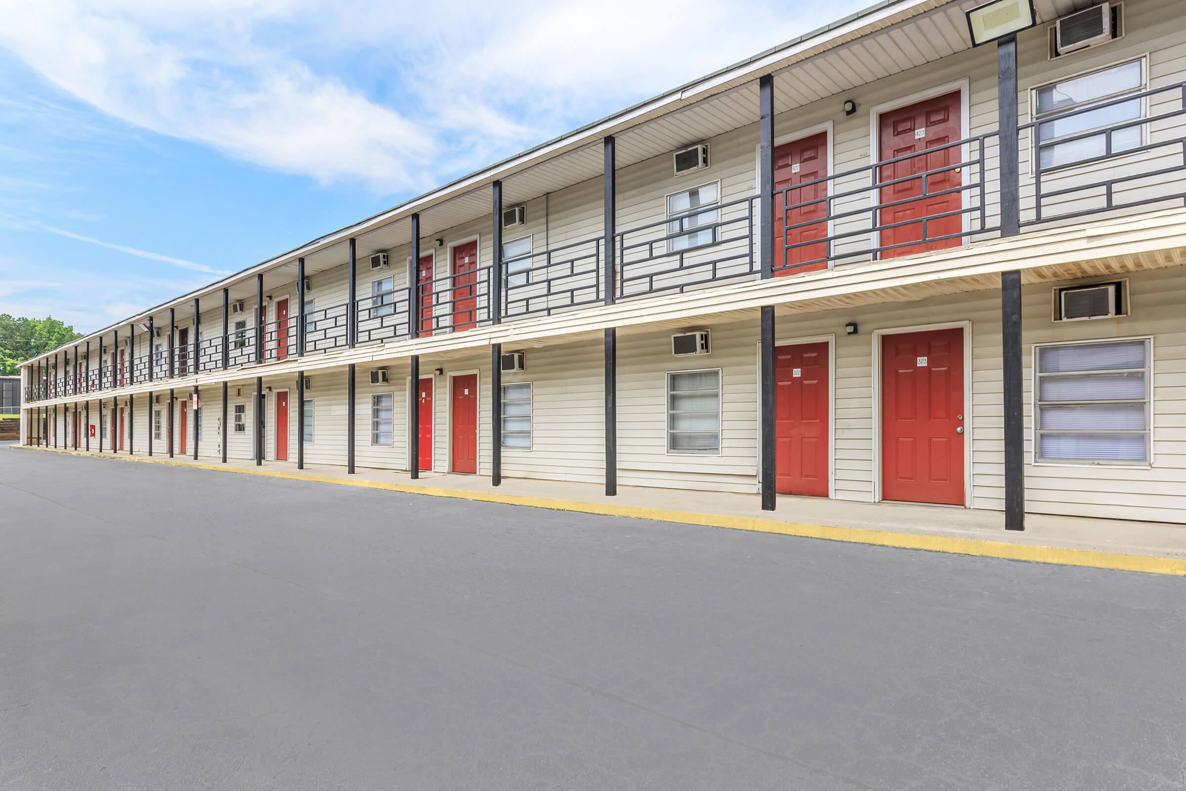 Row of motel rooms with red doors and black railings, set against a bright blue sky. The building features a simple, two-story design with multiple units, and a paved walkway in front. Air conditioning units are visible under windows, indicating a functional and welcoming space.