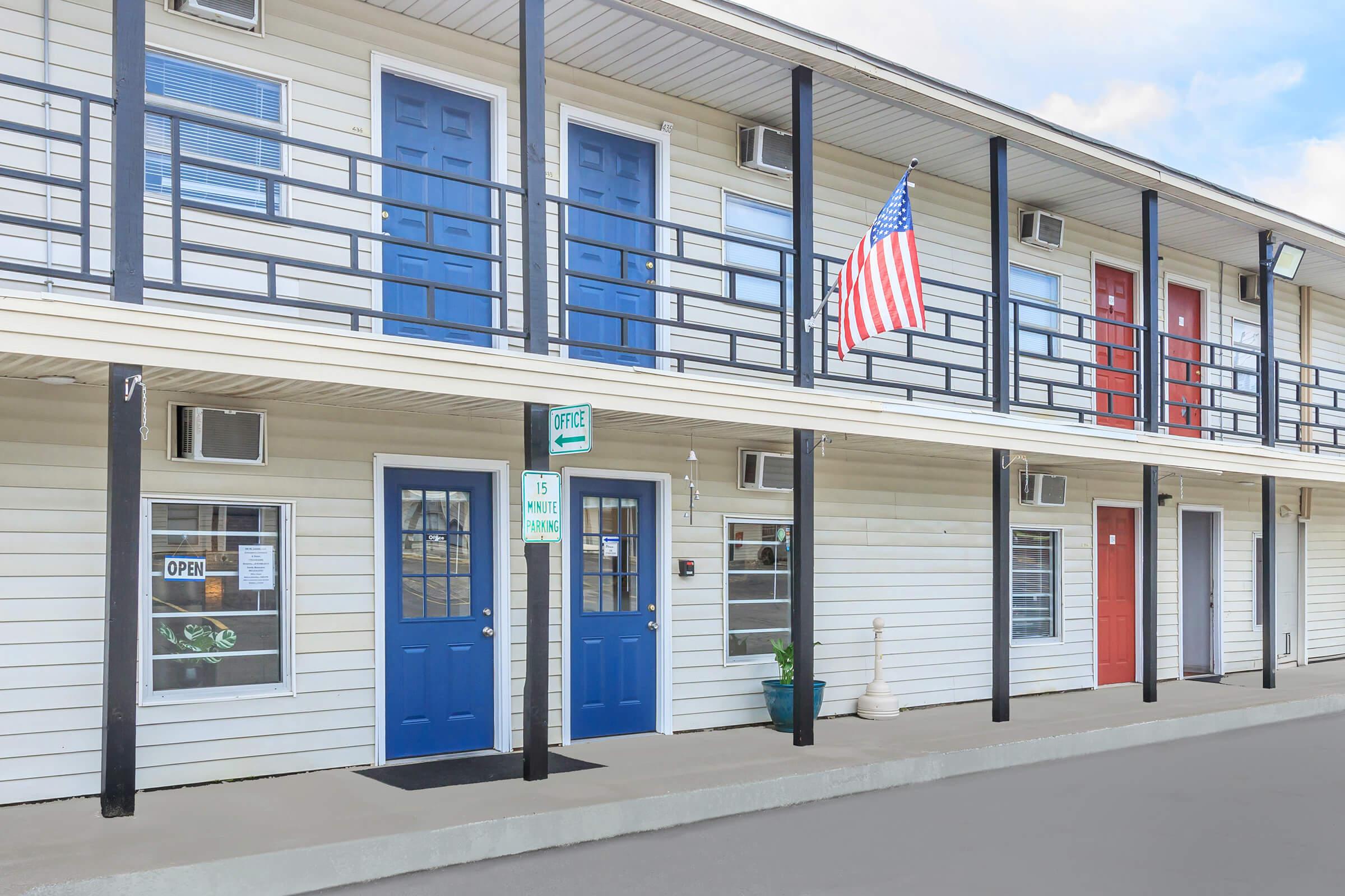 Exterior view of a motel with a two-story design, featuring blue and red doors, an American flag hanging from a balcony, and a green "OPEN" sign. The layout includes a paved area in front and several windows. The overall appearance is clean and welcoming, suitable for guests.