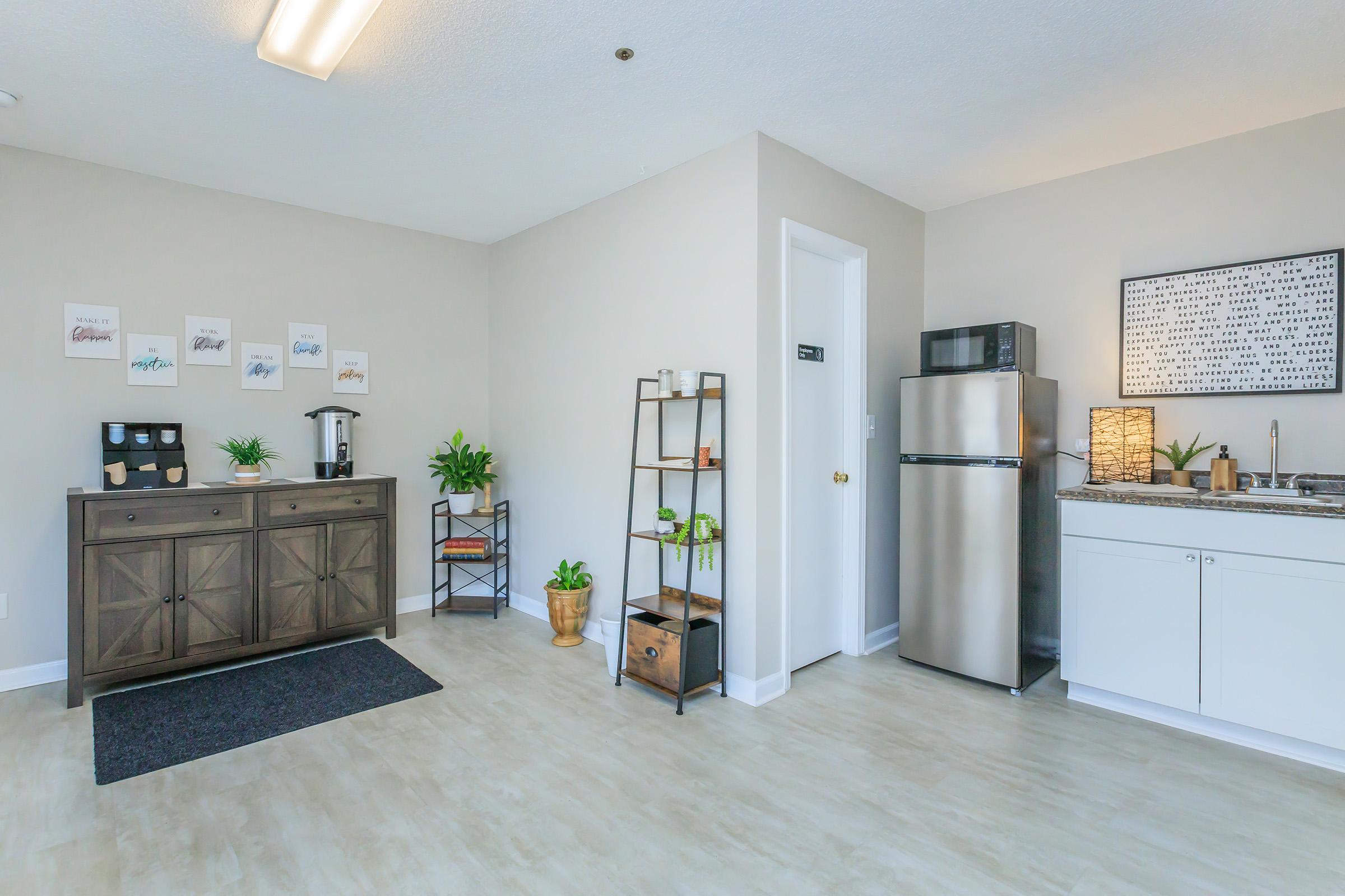A bright, modern kitchen area featuring a stainless steel refrigerator, a white cabinet, and a wooden sideboard displaying decorative items. There are plants, a coffee maker, and wall art with inspirational quotes. The room has neutral-colored walls and a warm, inviting atmosphere.