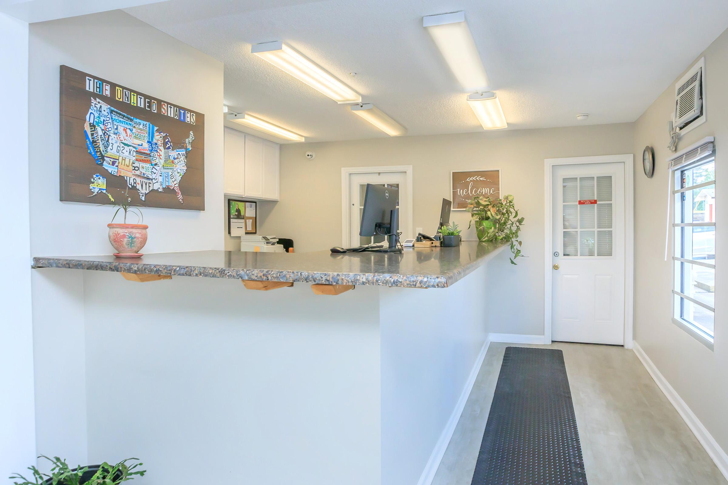 A bright and welcoming reception area featuring a long counter with a floral arrangement, a map art piece on the wall, and a monitor. Natural light coming from the windows enhances the inviting atmosphere. A welcome sign adds to the friendly vibe of the space.