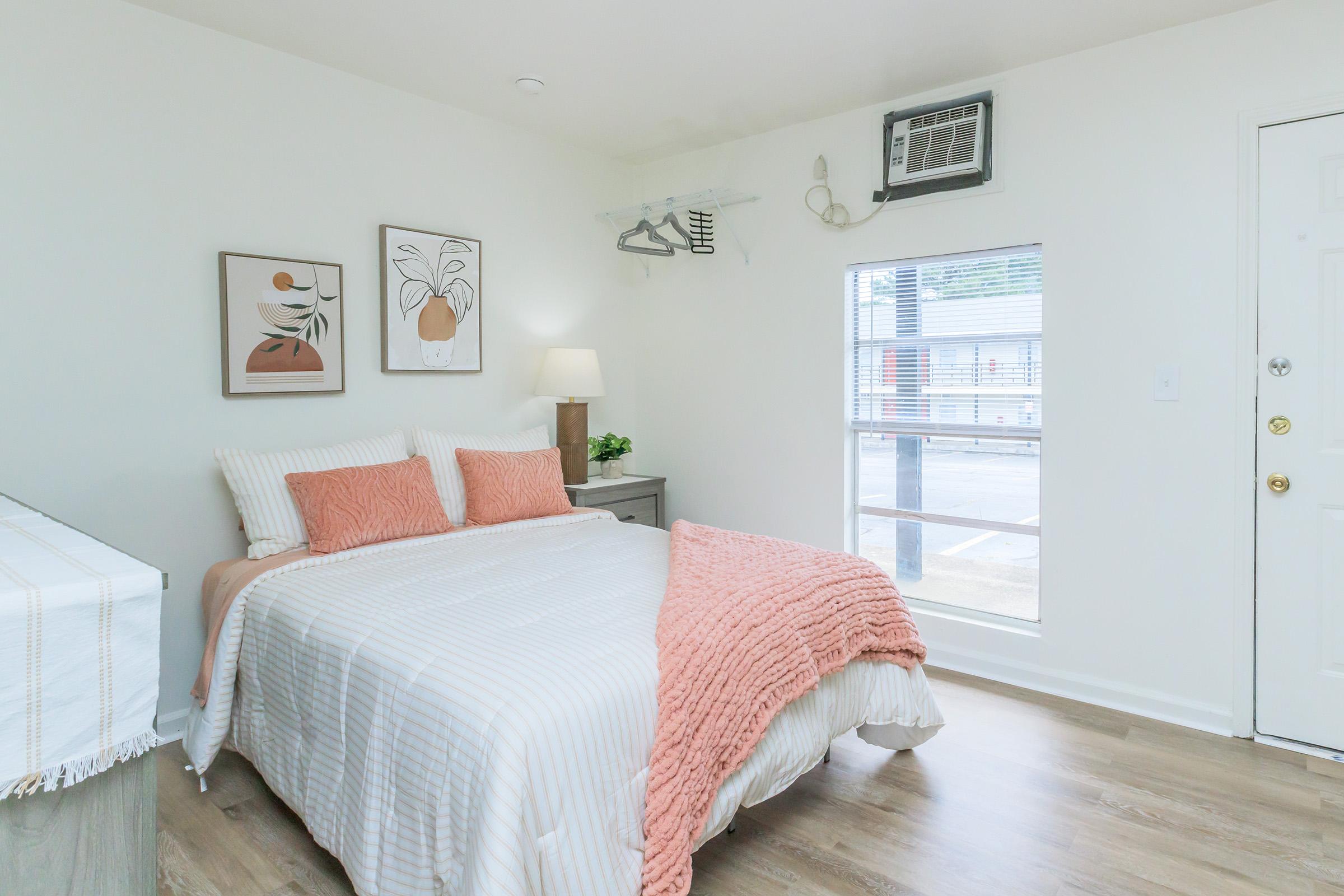 Cozy bedroom with a double bed featuring light-colored bedding and coral throw pillows. There's a bedside lamp on a dresser, and artwork on the walls. An air conditioning unit is mounted above the window, which lets in natural light. Wood flooring completes the inviting atmosphere.
