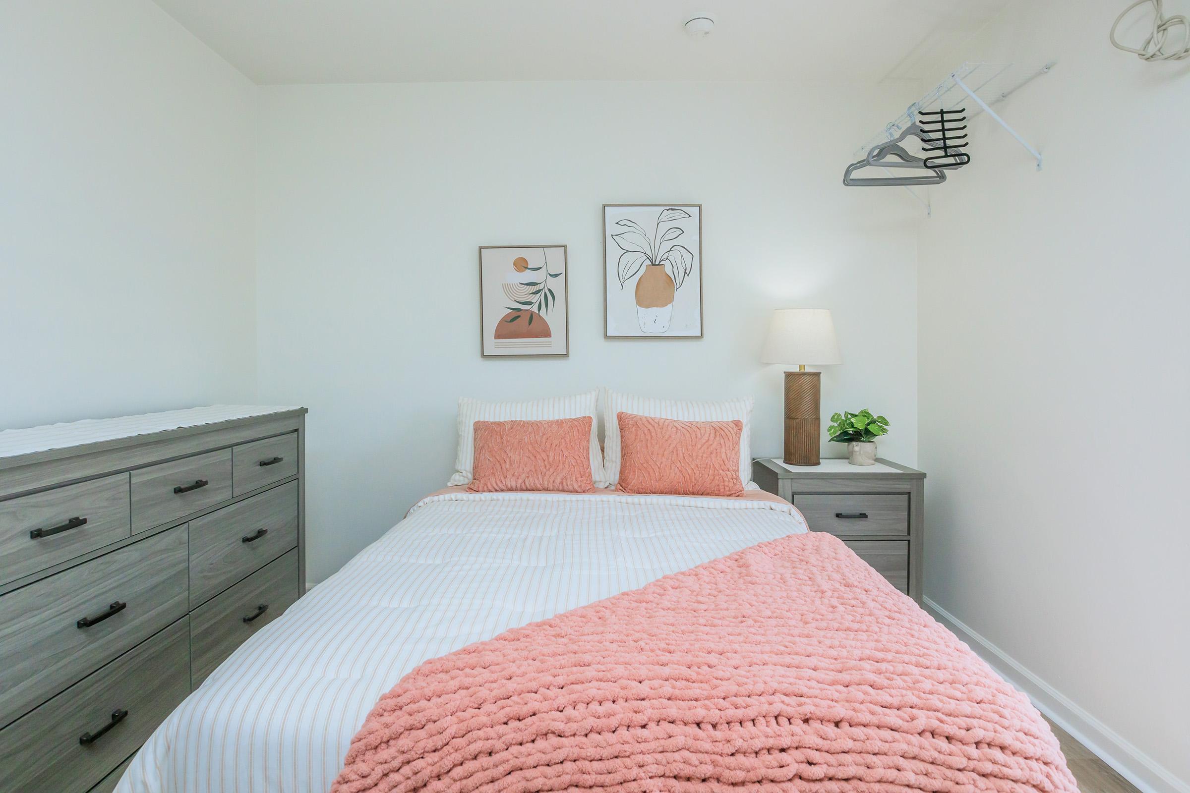 A cozy bedroom featuring a double bed with coral pillows and a textured pink throw blanket. On the wall, there are two framed artworks depicting plants. A bedside lamp and a small potted plant add a touch of decor, while a light-colored dresser is positioned next to the bed. The walls are painted white, creating a bright atmosphere.