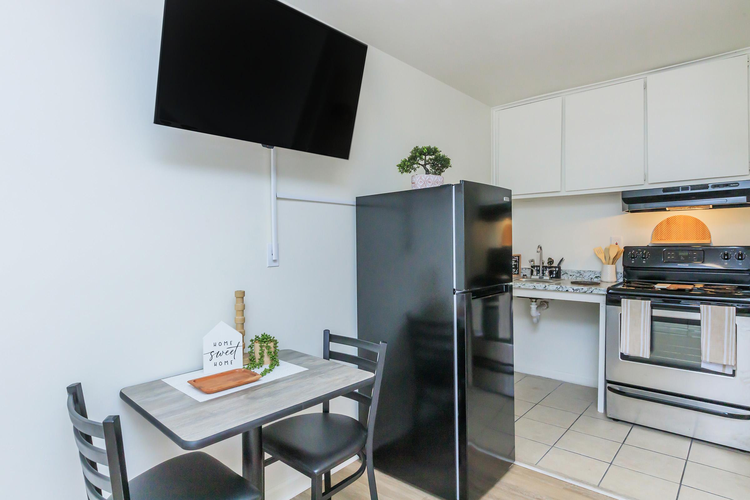 A modern kitchen featuring a black refrigerator, an oven and stovetop, and a flat-screen TV mounted on the wall. There is a small dining table with two black chairs and a decorative sign. The kitchen has white cabinets and light-colored tile flooring, creating a clean and contemporary look.