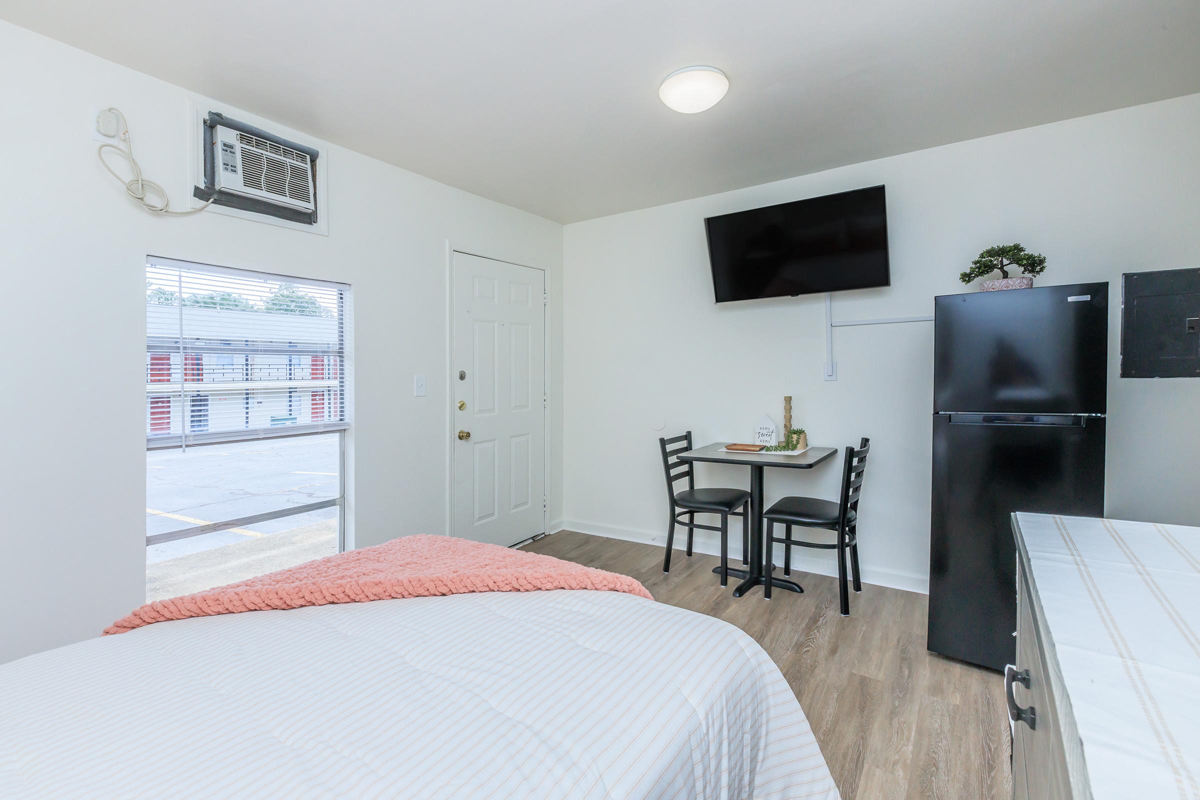 A cozy interior of a small room featuring a bed with a coral throw, a kitchenette with a refrigerator, a black table with two chairs, a mounted TV on the wall, and a window with natural light. An air conditioning unit is visible above the door.