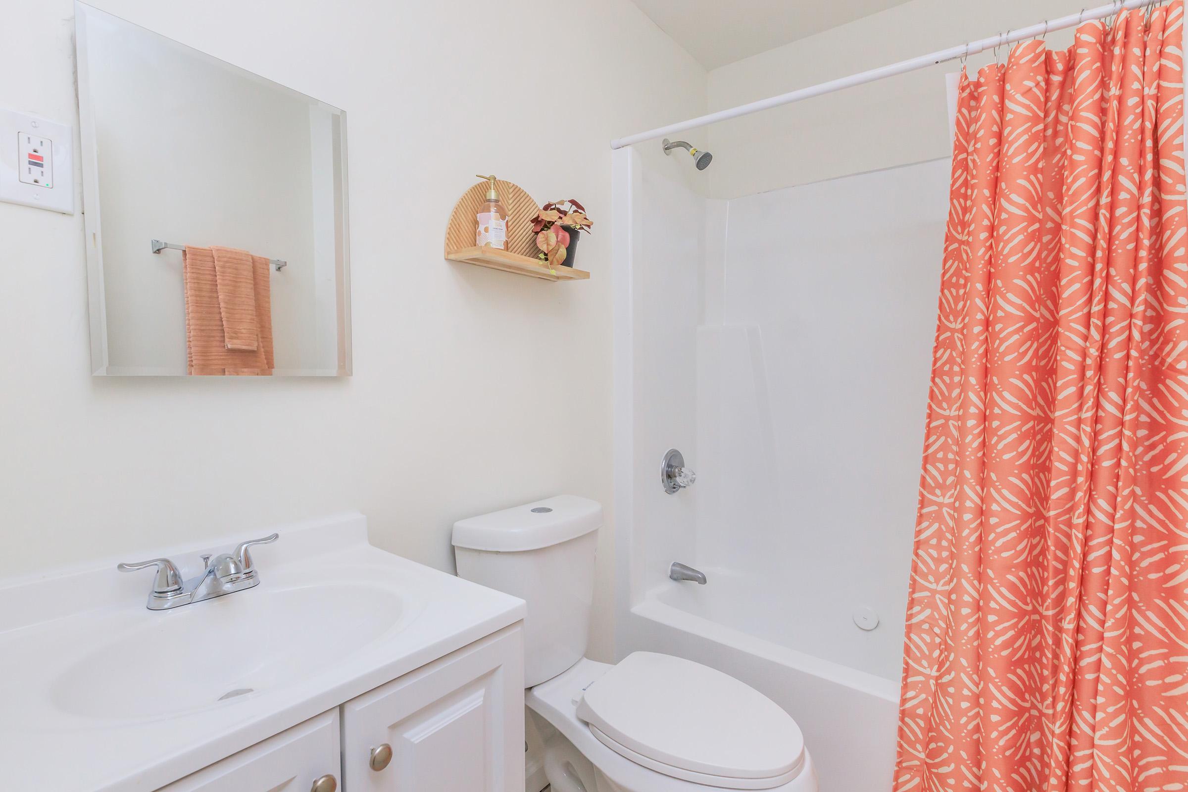 A bright bathroom featuring a white sink and cabinet, a white toilet, and a bathtub with a shower curtain in coral with a floral pattern. There’s a small shelf with decorative items and a mirror above the sink. A beige towel hangs on a hook next to the mirror. The walls are painted a light color, creating an airy feel.