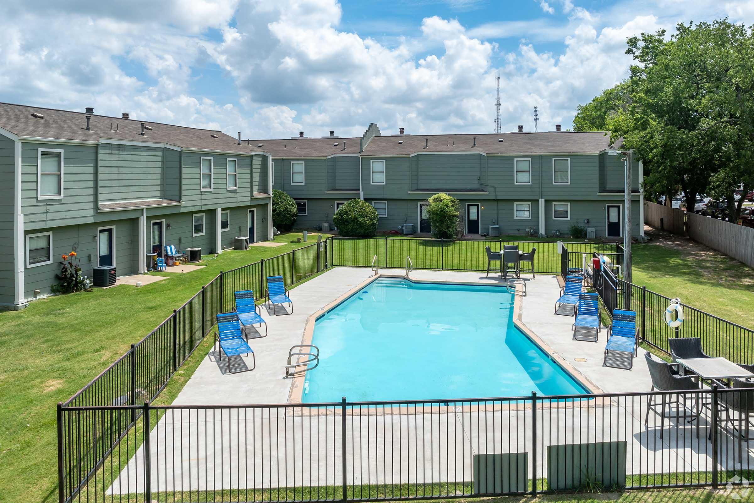 A clear blue swimming pool surrounded by a black wrought iron fence, with several blue lounge chairs positioned around it. In the background, multiple green apartment buildings are visible under a partly cloudy sky. Lush grass and trees are present, creating a welcoming outdoor environment.