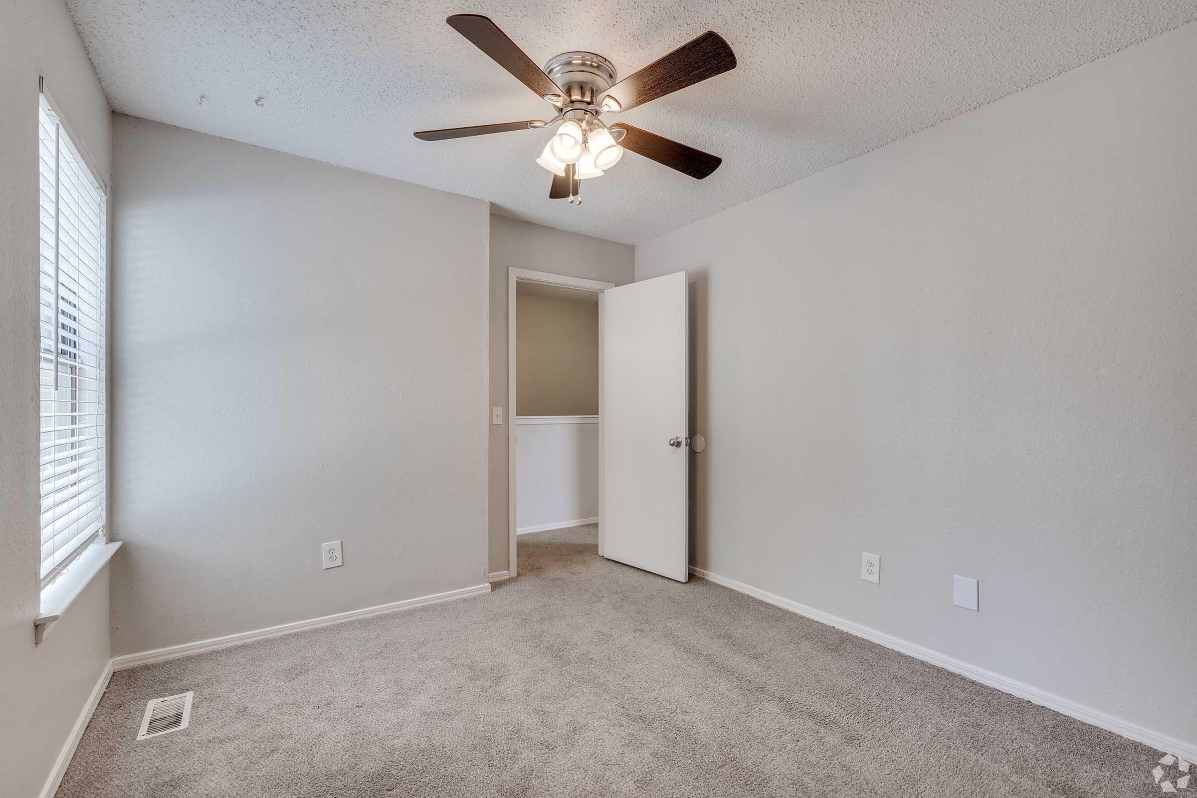 A small, empty room featuring beige carpet and light gray walls. There's a window with white blinds on one side, letting in natural light. A ceiling fan with four lights hangs above, and a white door is slightly ajar leading to another room. The overall atmosphere is bright and inviting.