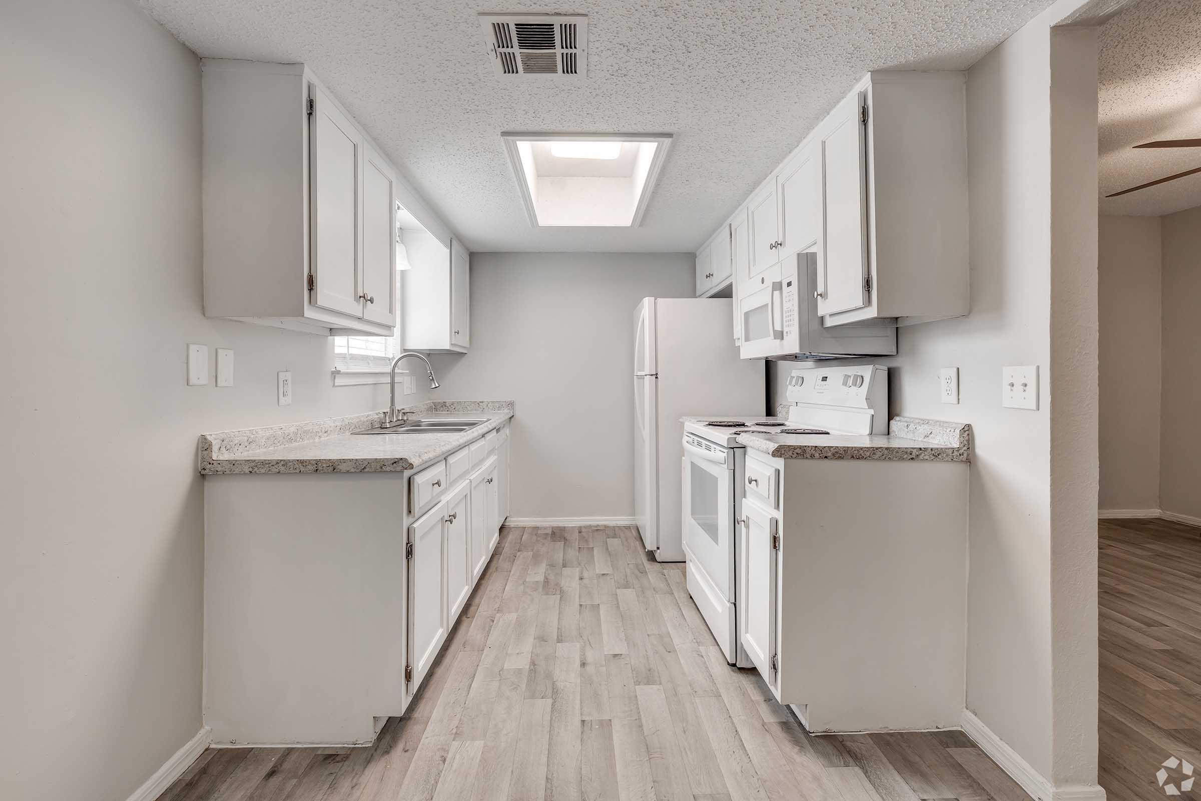 Modern kitchen interior featuring white cabinets, a sink, and stainless steel appliances. A skylight provides natural light, and light-colored wood laminate flooring enhances the space. The layout is open and functional, ideal for cooking and entertaining.